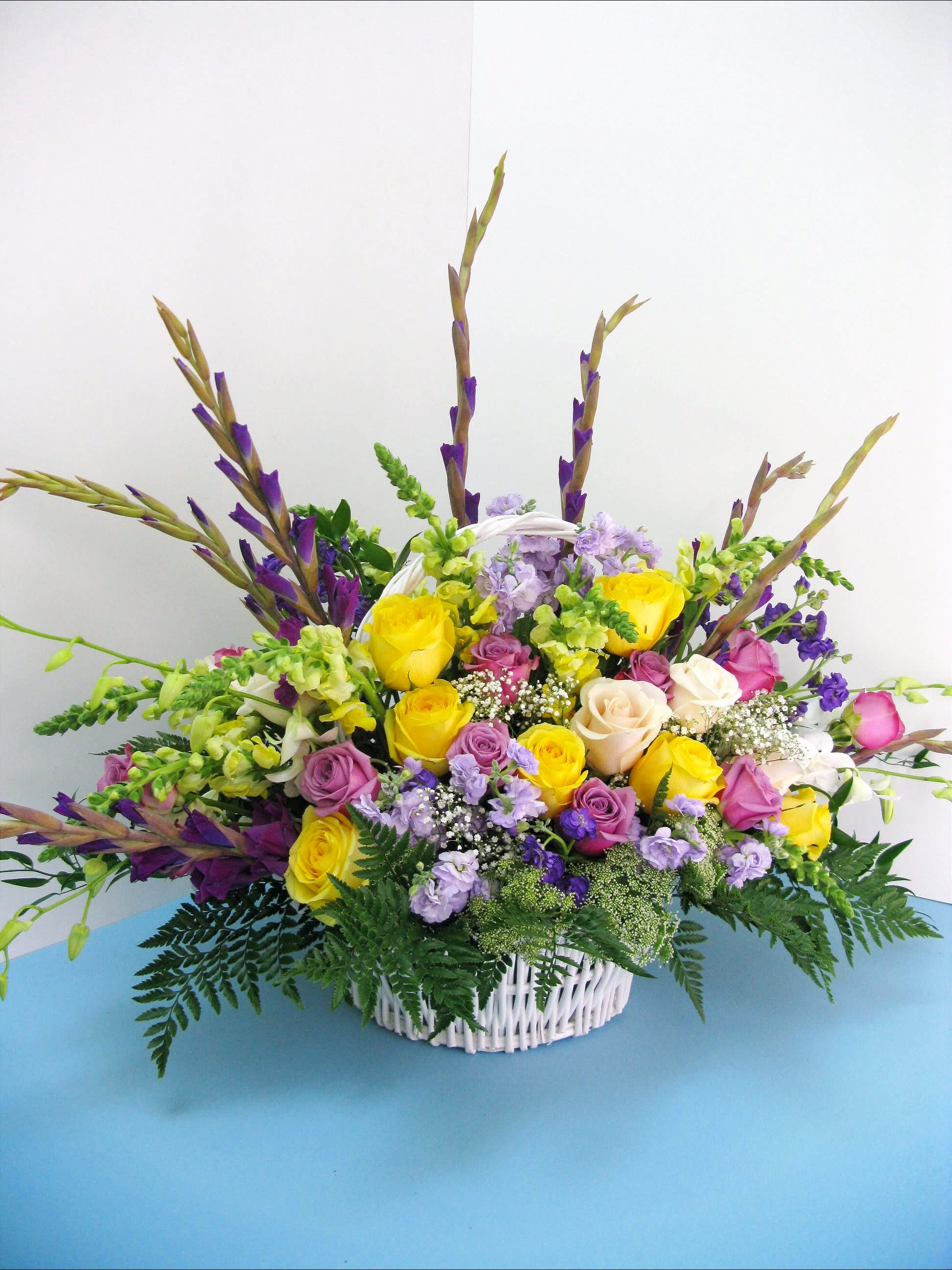 Tall mixed arrangement of yellow and pink roses with purple accents in a white wicker basket