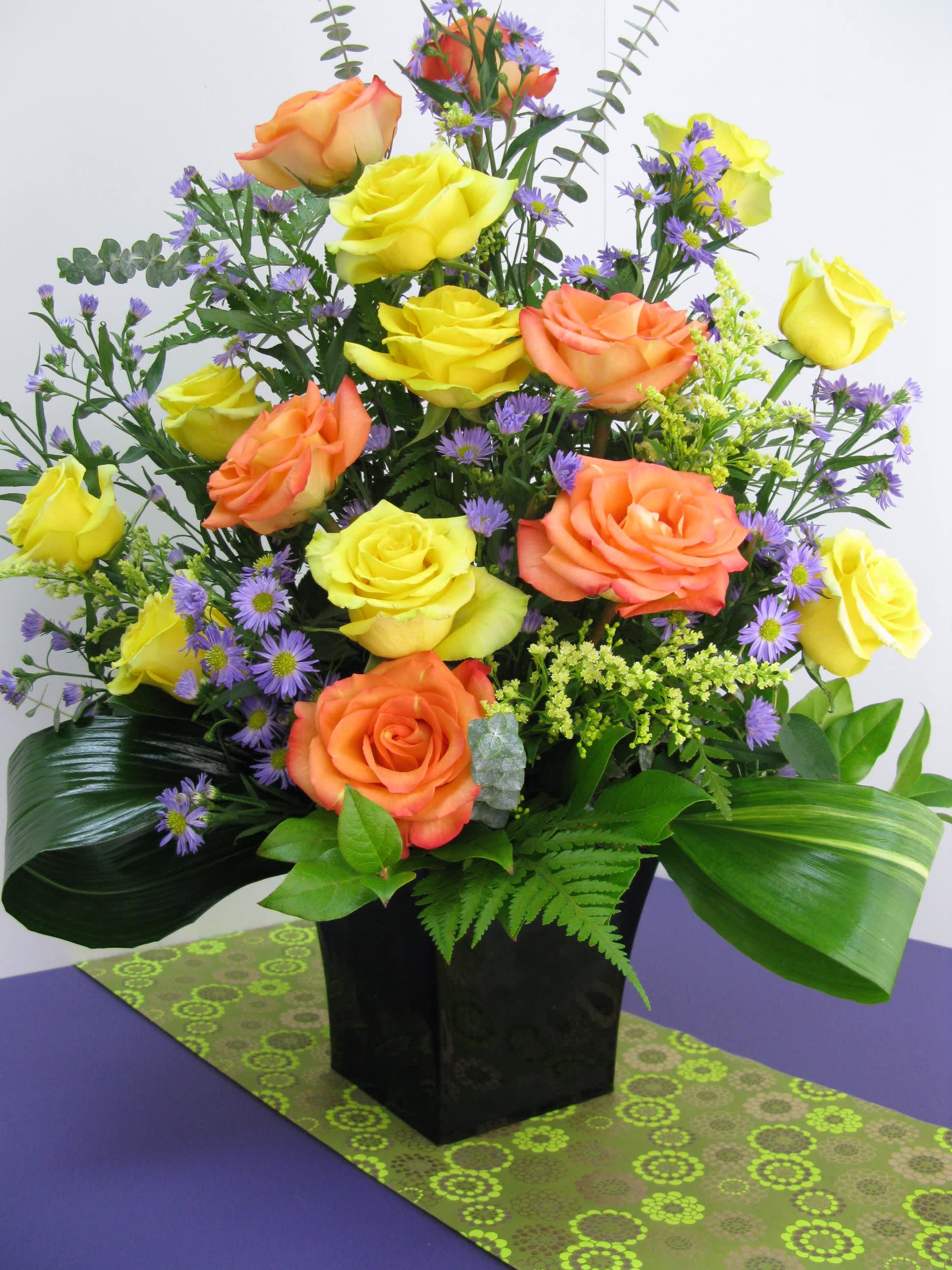 Tall arrangement of yellow and orange roses with small purple flowers in a black cube vase