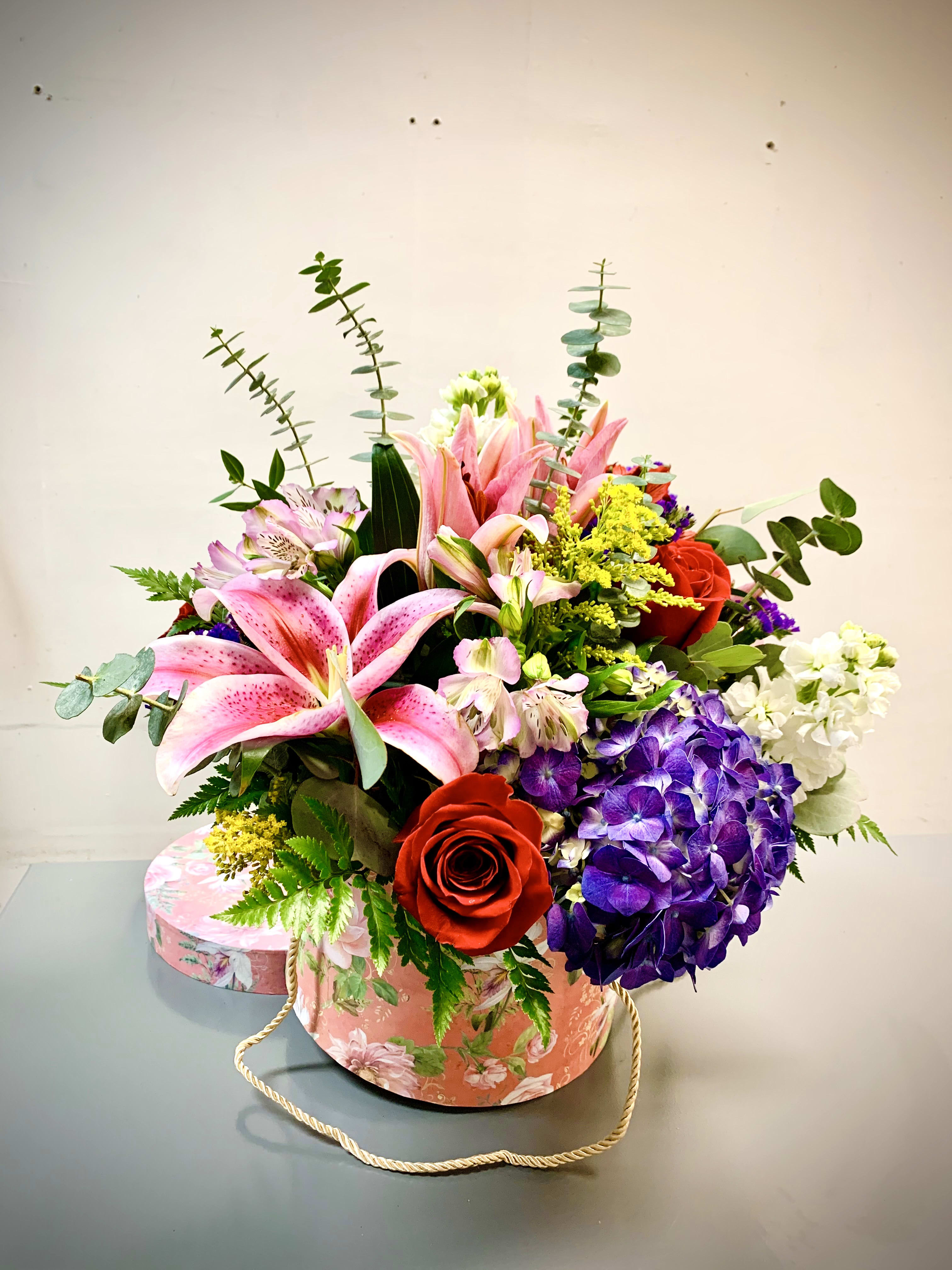 Mixed bouquet with pink lilies, red roses, purple hydrangea, and white blooms in a floral hatbox