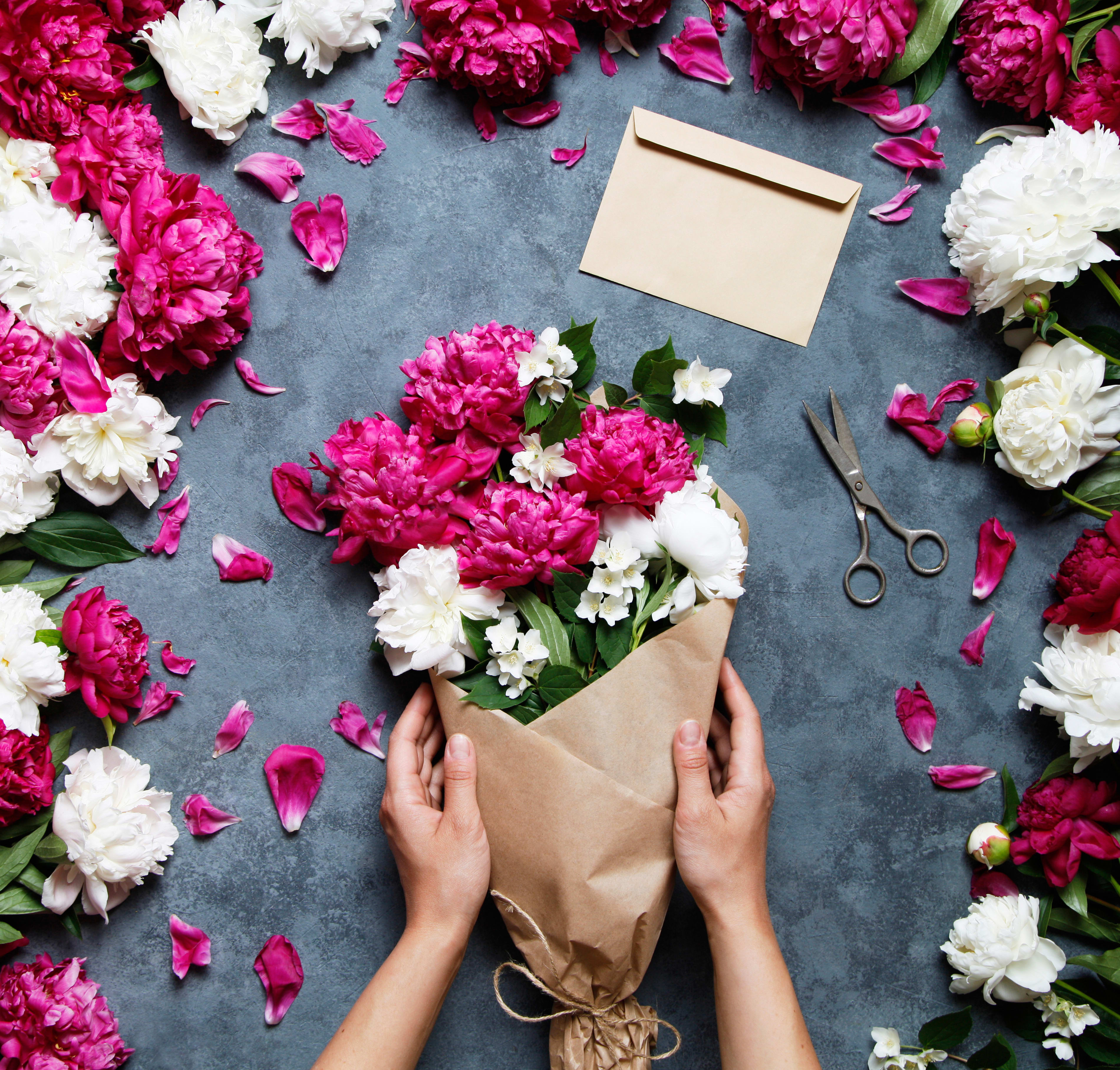 Hand-held bouquet of pink and white peonies in brown paper with an envelope and scissors nearby