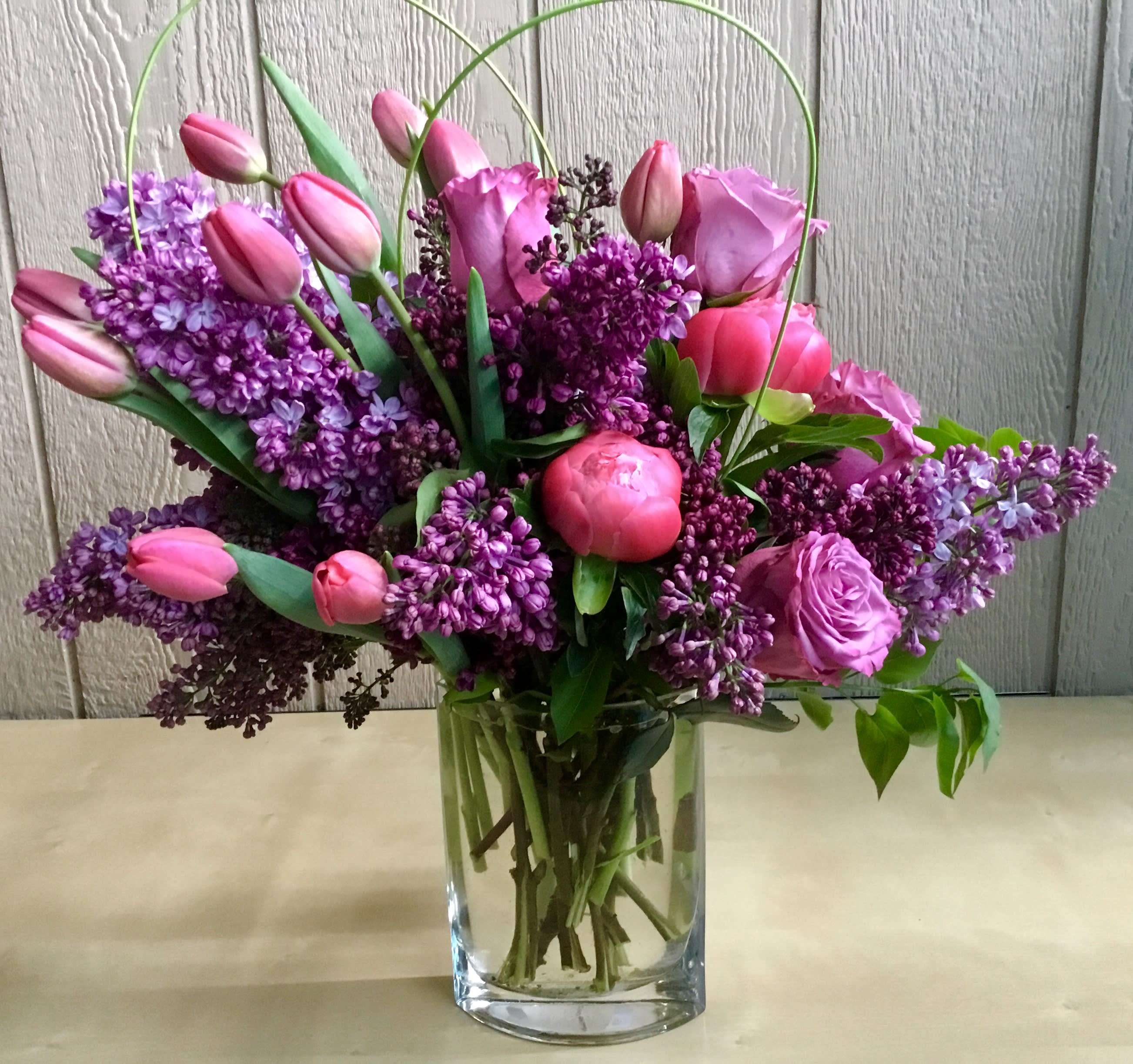 Pink tulips, roses, peonies, and purple lilacs arranged in a clear glass vase