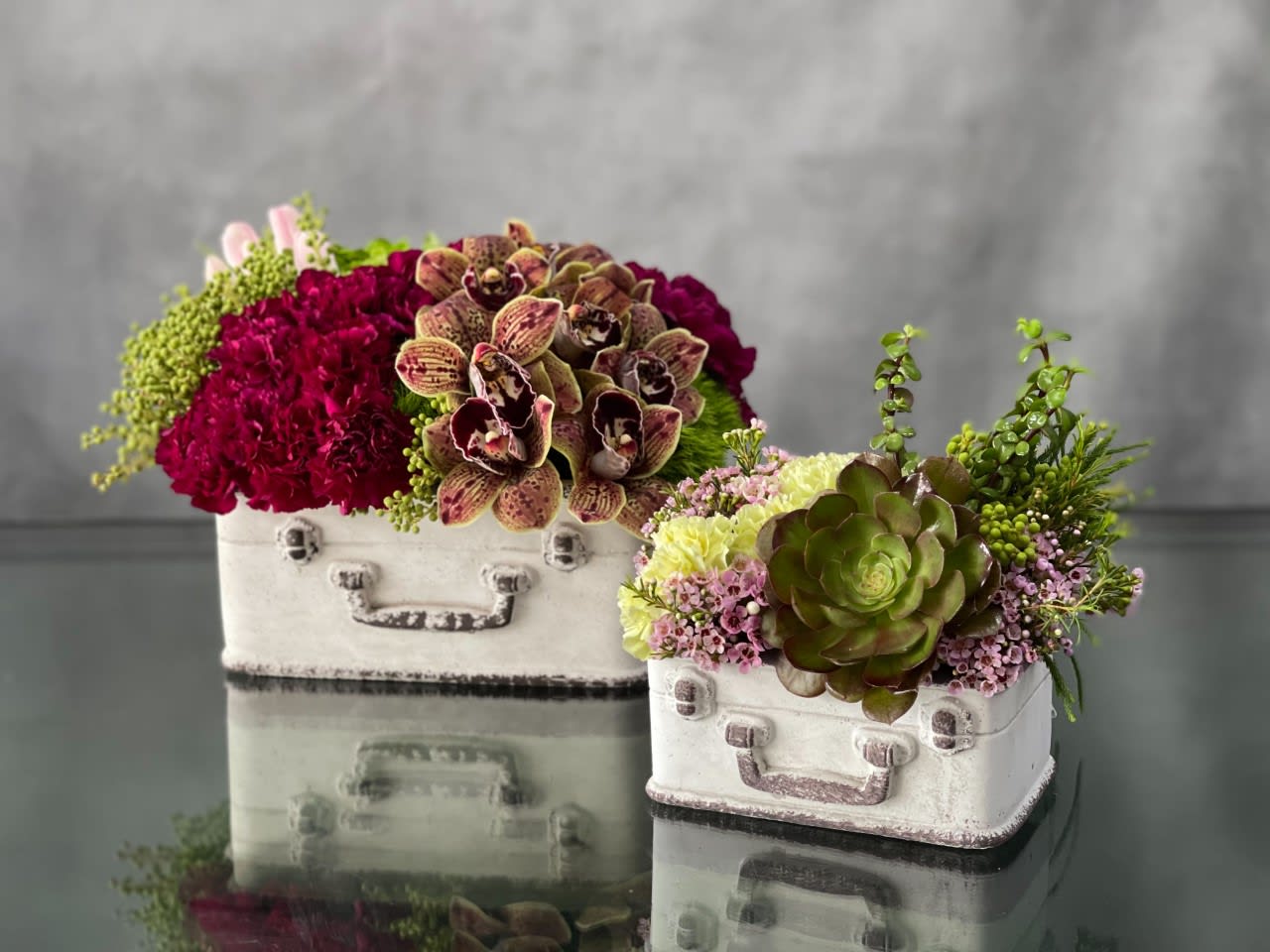 Traveling Blooms - This whimsical set of suitcase planters is a great gift to someone who is about to travel or back from traveling. The larger planter is filled with spotted cymbidium orchids, red velvet carnations, and pink dutch tulips. The smaller planter has succulents, wax flowers, and cream carnations. What a fun and unique gift !  Large planter: 15x12 Small planter : 9x8 