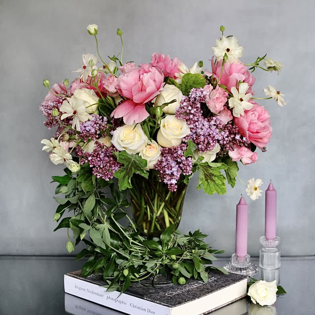 Ophelia  - "Ophelia" is a true notion of delicacy. This arrangement consists of Lilacs, Peonies, Butterfly Ranunculus and seasonal florals and greens . Greens that wrap around the base of a medium length glass pedestal vase. Beautifully arranged for that special someone in your life.   Measurement:  Vase: 12 x 8 Arrangement: 23 x 19