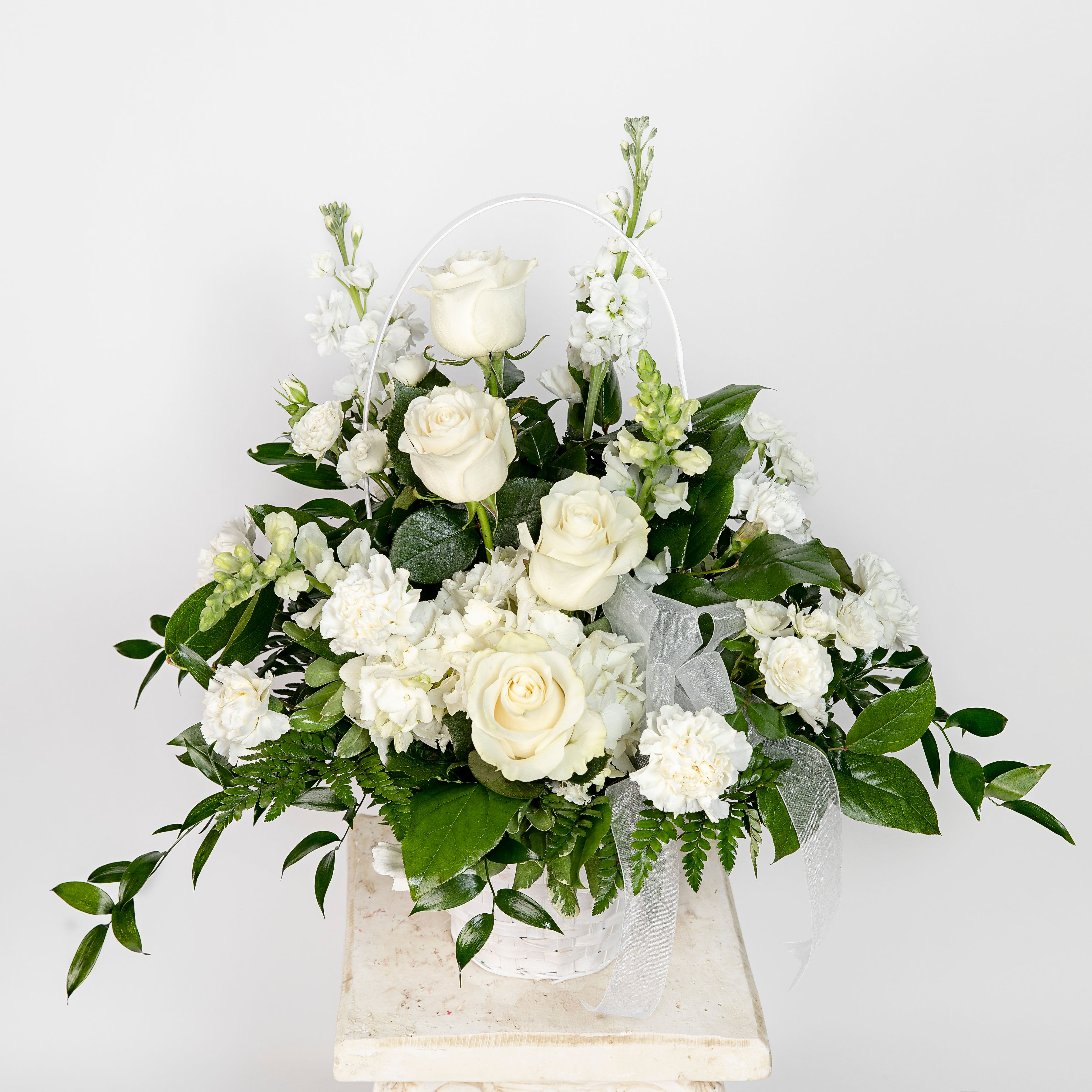 White floral basket with roses and carnations arranged with lush foliage and a sheer ribbon bow.