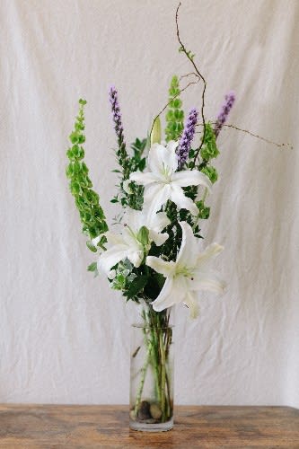 Tall white lily arrangement with green and purple spikes in a glass vase