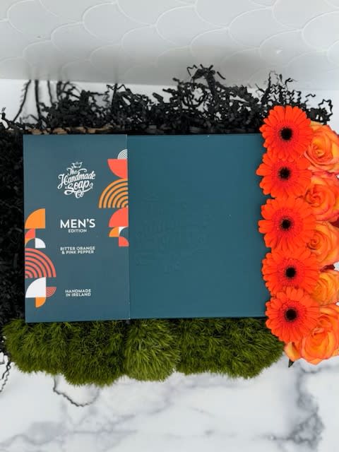 Men's soap gift box with bright orange gerbera daisies and roses, green moss and black filler in a tray