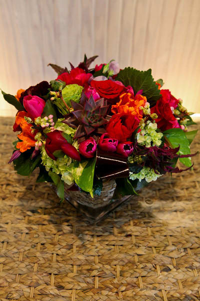 Low mixed arrangement of red roses, pink tulips, green hydrangeas, and a central succulent in a glass cube vase.
