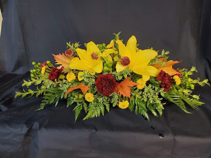 Leaves and Lilies long and low centerpiece - A fresh mix of Fall colored leaves and beautiful fall colored lilies. This centerpiece is shaped perfectly to encourage conversations as you will never have to try and see around it.