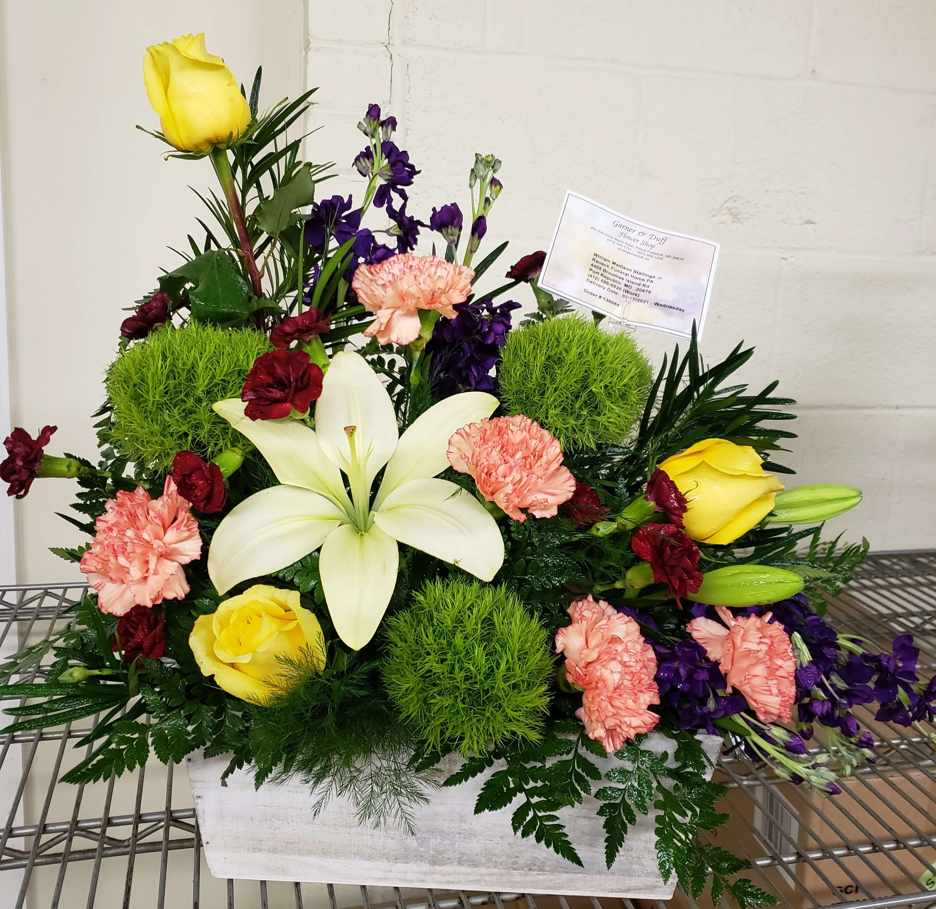 Mixed floral arrangement with yellow roses, white lilies, and pink carnations in a white container