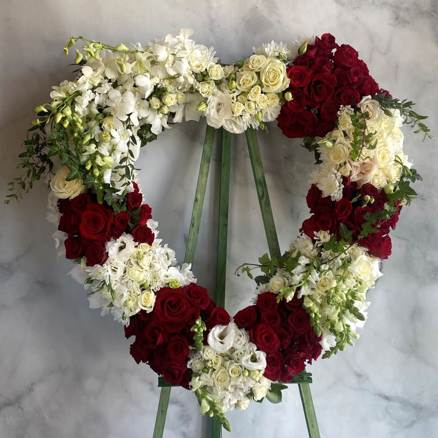 Flowers Fill Your Heart Wreath II - This beautiful open heart wreath is a heartfelt tribute to a loved one. Adorned with a mix of elegant red and white roses, delicate dendrobium orchids, and lisianthus, this arrangement is a symbol of love and affection. The green ivy provides a natural touch to this stunning display. Measuring approximately 27 inches in diameter, this open heart wreath is the perfect tribute to honor the memory of a dear friend or family member.