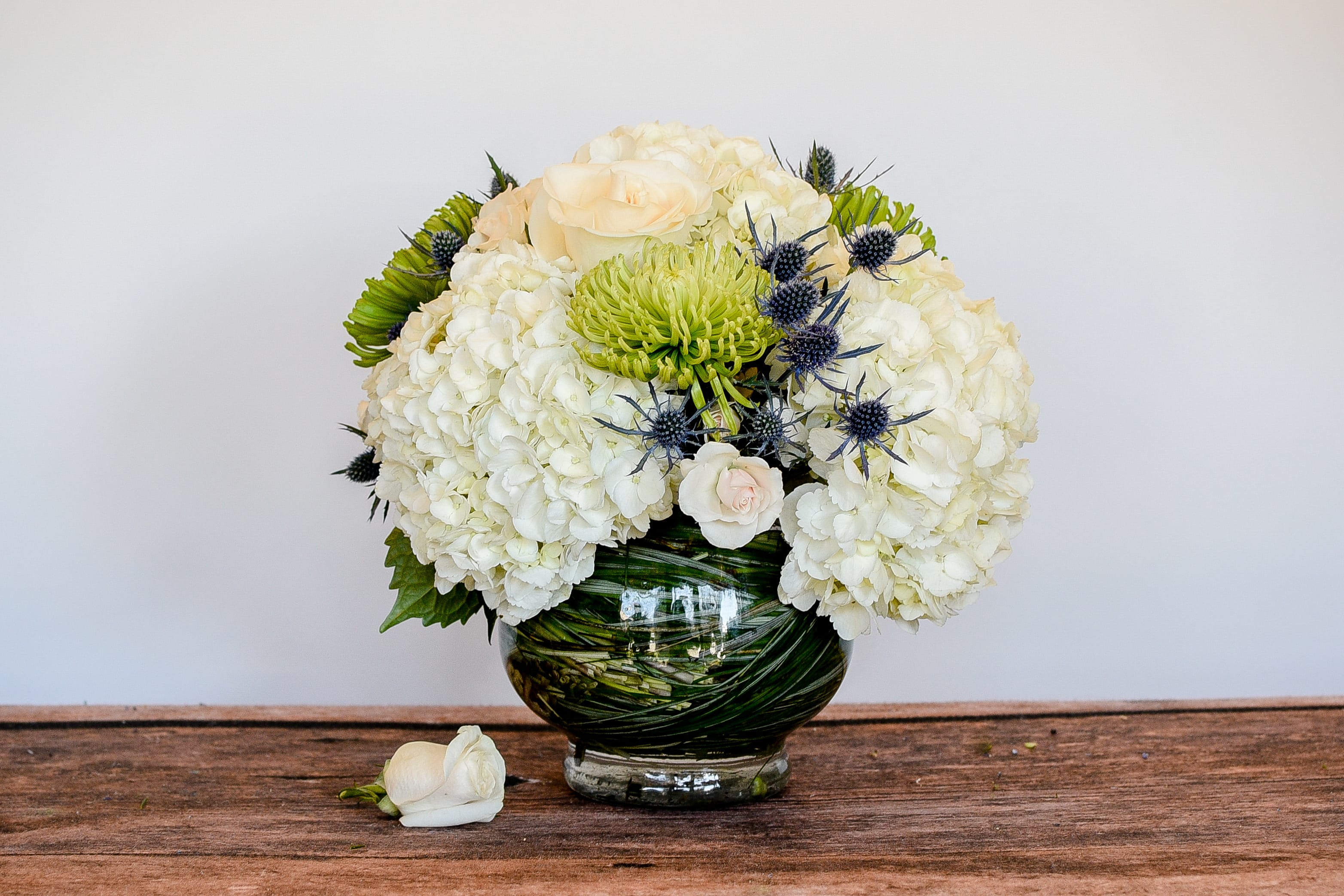 Low round arrangement of white hydrangeas, roses, and green mums in a leaf-wrapped glass bowl vase