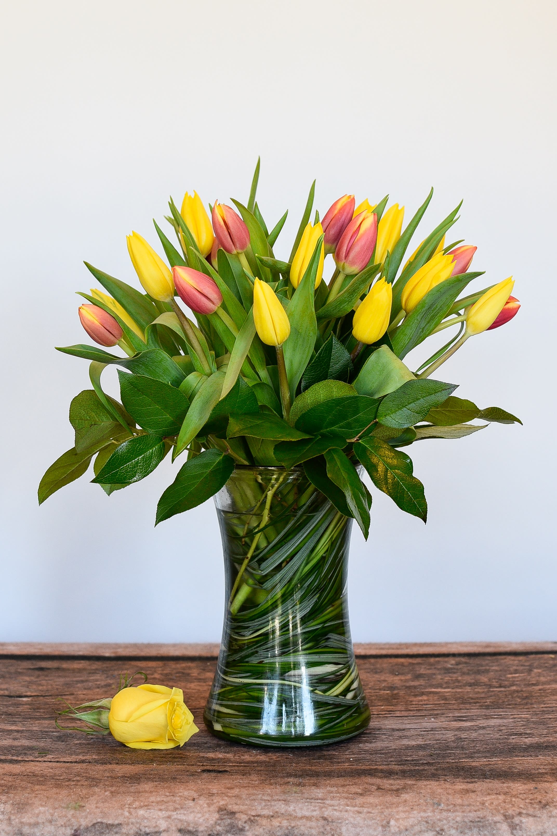 Yellow and pink tulips in a clear glass vase with a single yellow rose lying beside it