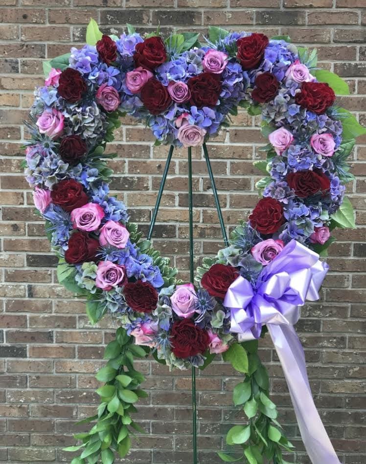 Heart-shaped standing wreath of red and pink roses with purple hydrangeas and a lavender bow on an easel.