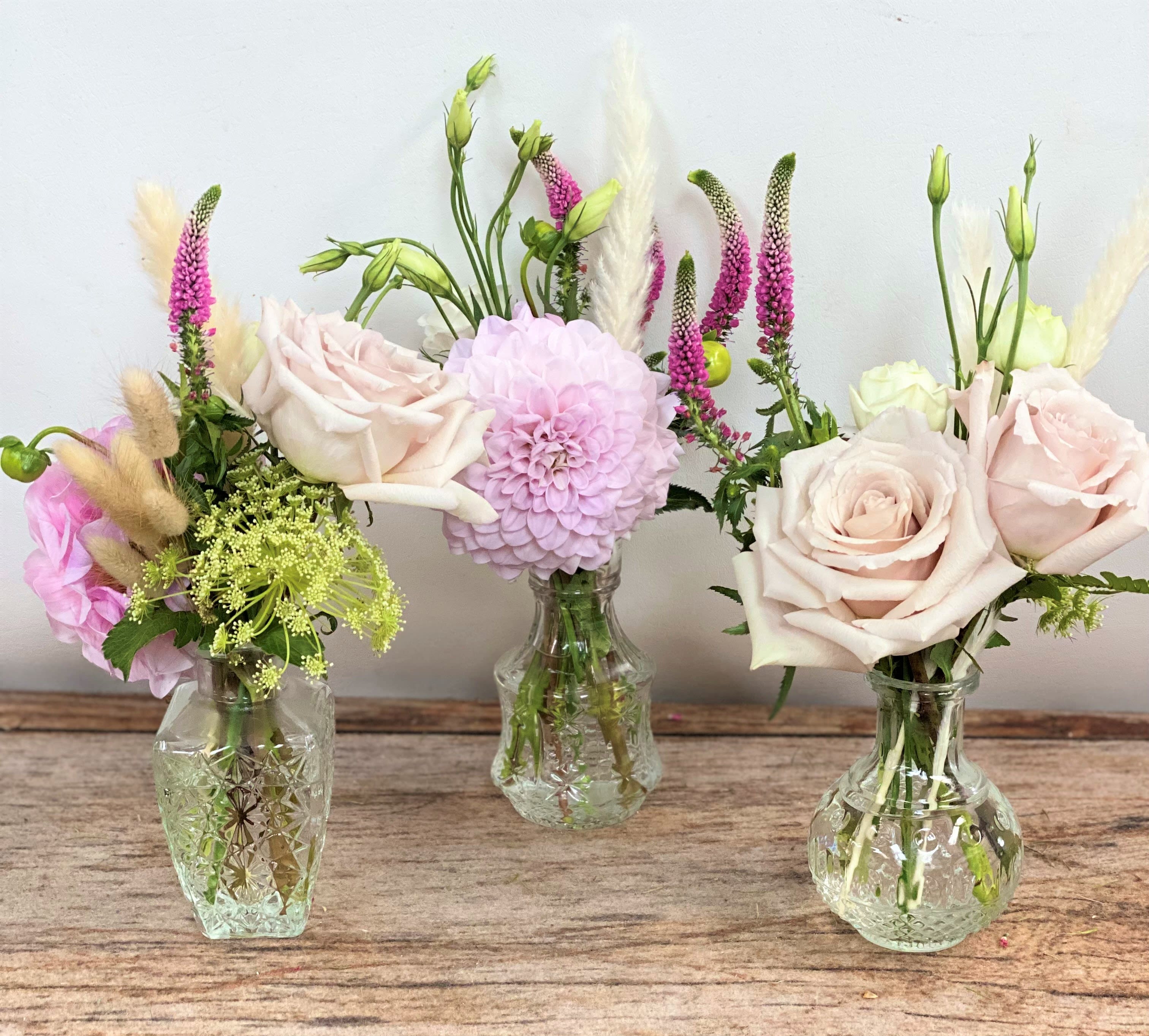 Trio of small glass bud vases with blush roses, a pink dahlia, and delicate accent flowers.