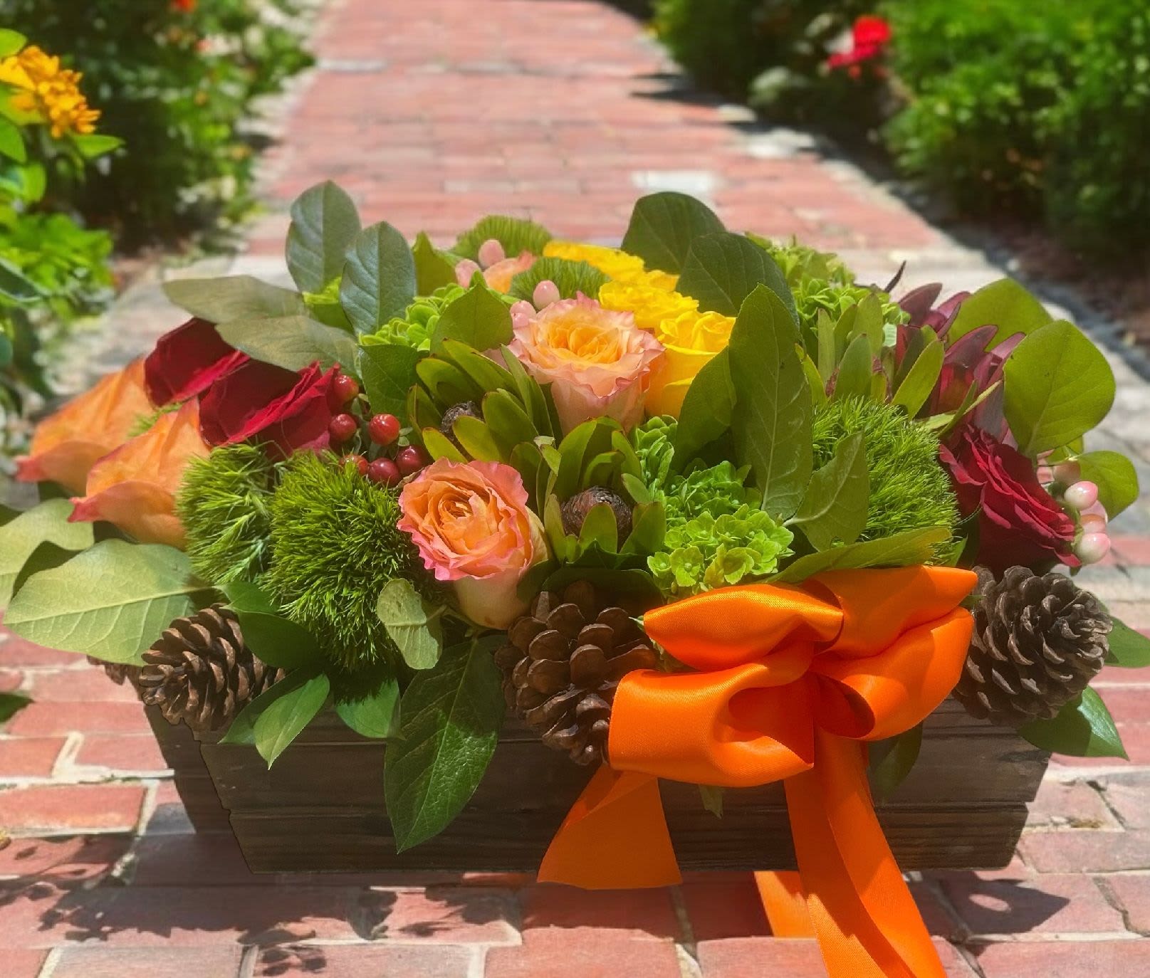 Autumn Bliss - Autumn Bliss | Wilmington, Delaware Florist  Celebrate the vibrant colors of the season with Autumn Bliss, a rustic yet elegant floral arrangement handcrafted by Petals Flowers and Fine Gifts in Wilmington, Delaware. This striking design features lush green trichelium, golden yellow roses, warm orange roses, deep red roses, and bold red protea. Accented with natural pine cones and tied with an elegant orange satin ribbon, the flowers are beautifully arranged in a long rectangular wooden container for a centerpiece full of seasonal charm. Perfect as a Thanksgiving centerpiece, fall gathering décor, or a thoughtful gift, Autumn Bliss blends rich color and natural texture to create a warm, inviting design. Whether displayed on a dining table, console, or office reception area, this arrangement adds a touch of autumn elegance to any space. As your trusted local florist in Wilmington, Delaware, Petals Flowers and Fine Gifts is proud to design luxury floral arrangements with fresh, premium blooms and seasonal accents. We offer same-day, tax-free flower delivery throughout Wilmington, Hockessin, Newark, Chadds Ford, Glen Mills, Kennett Square, and surrounding communities—ensuring your flowers arrive fresh and beautiful every time. Why Choose Petals Flowers and Fine Gifts? Unique, seasonal floral arrangements with luxury blooms Family-owned Wilmington, Delaware florist offering personal service Same-day, tax-free flower delivery to homes, offices, and event venues