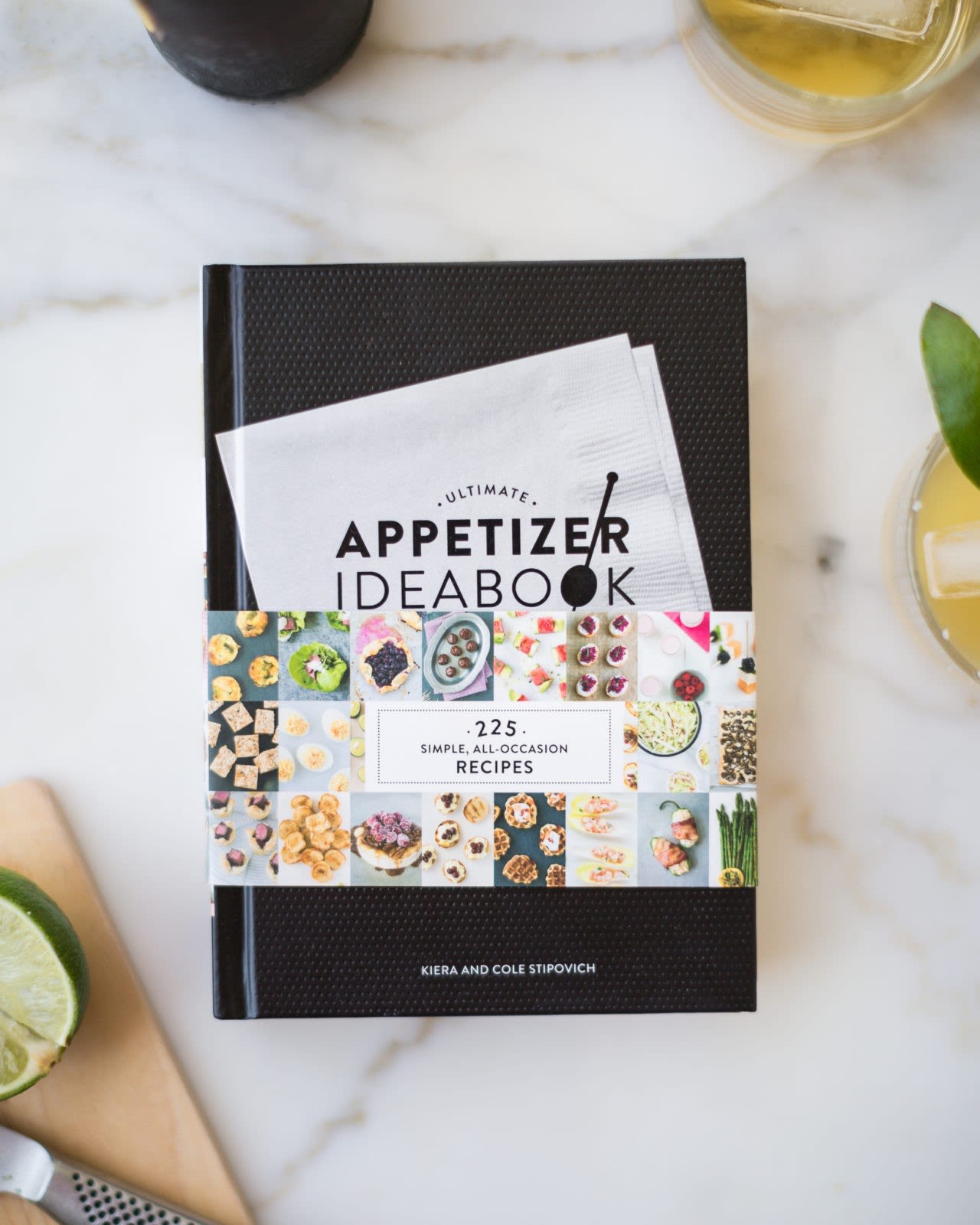 A cookbook on a marble surface with glasses of drink around it