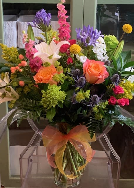 Bright mixed bouquet with orange roses, white lilies, and pink blooms in a clear vase with an orange bow