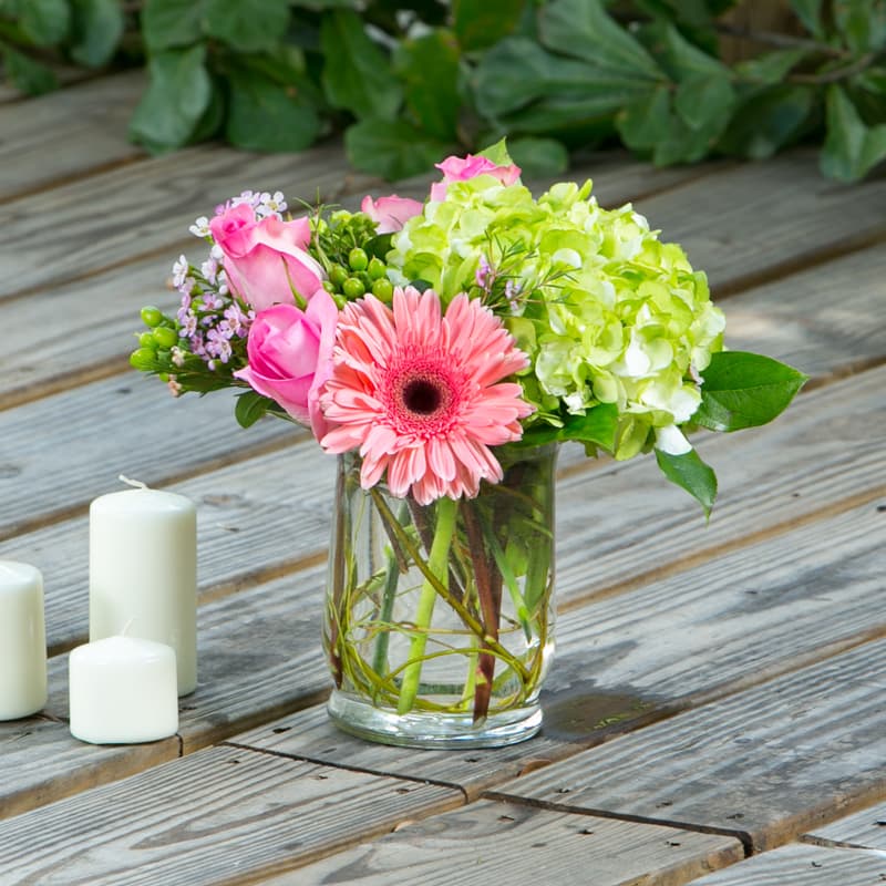 Pink roses and a gerbera daisy in a glass vase with candles nearby