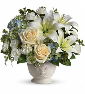 Ivory lilies, cream roses, and white carnations arranged with blue hydrangea in a white ceramic urn vase