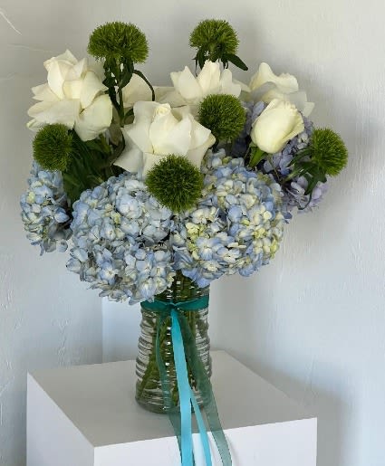 White roses and blue hydrangeas in a glass vase with green pom-pom accents