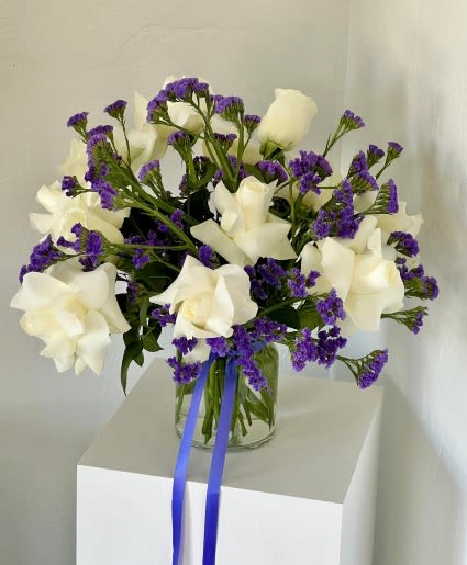 White roses and purple filler flowers in a clear glass vase with blue ribbon