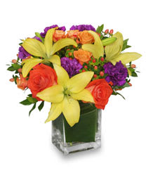 Bouquet of yellow lilies, orange roses, and purple flowers in a glass vase