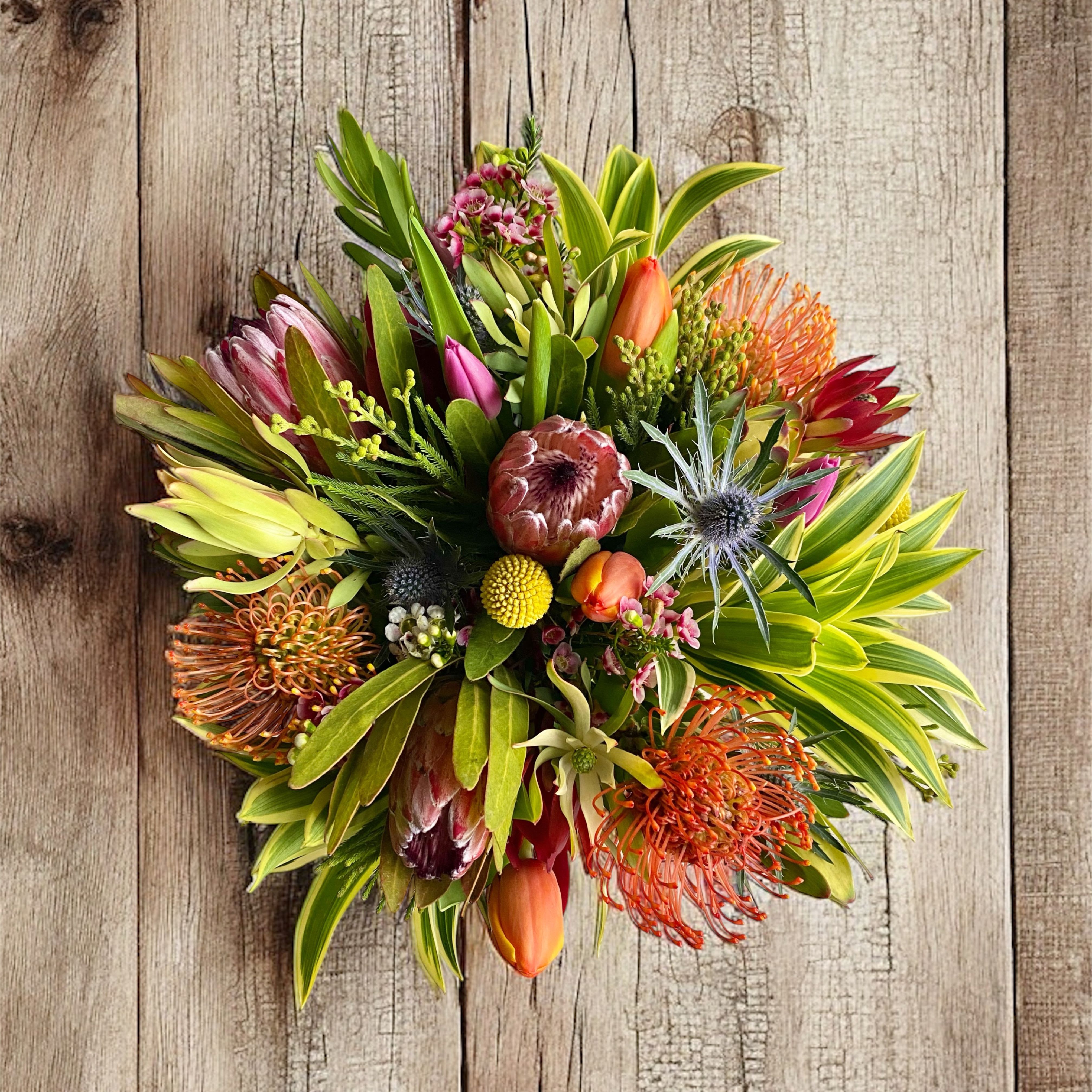 Round tropical bouquet with protea, pincushion blooms, and orange tulips on a wood background