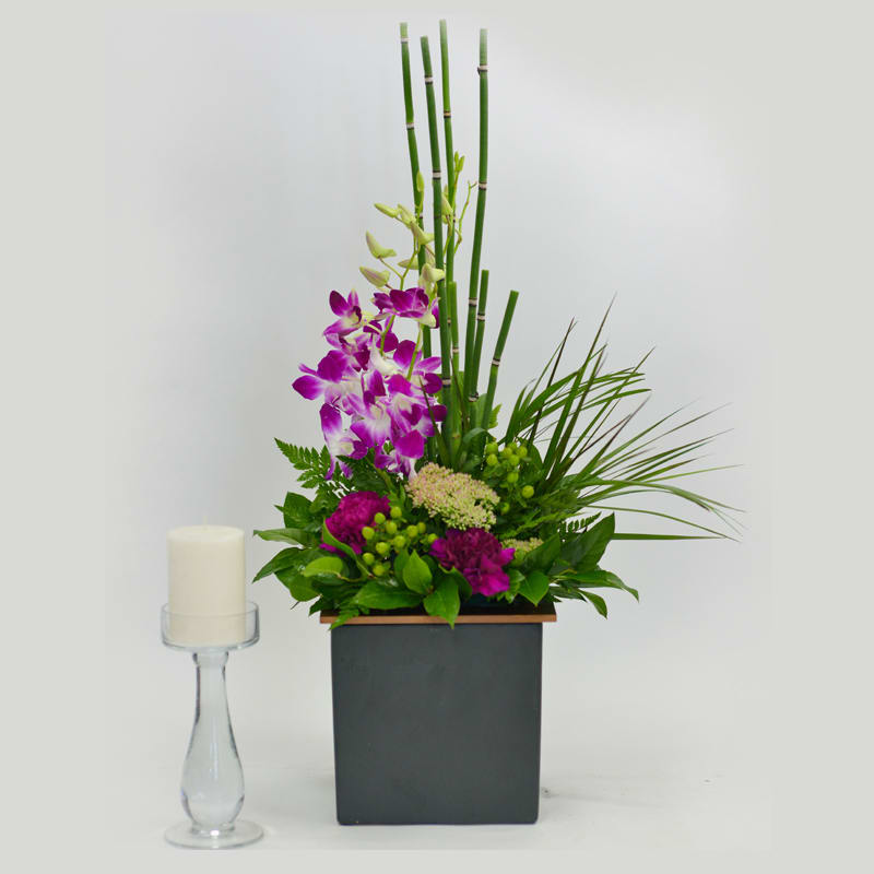 Tall orchid arrangement in a square black vase beside a white candle on a glass holder