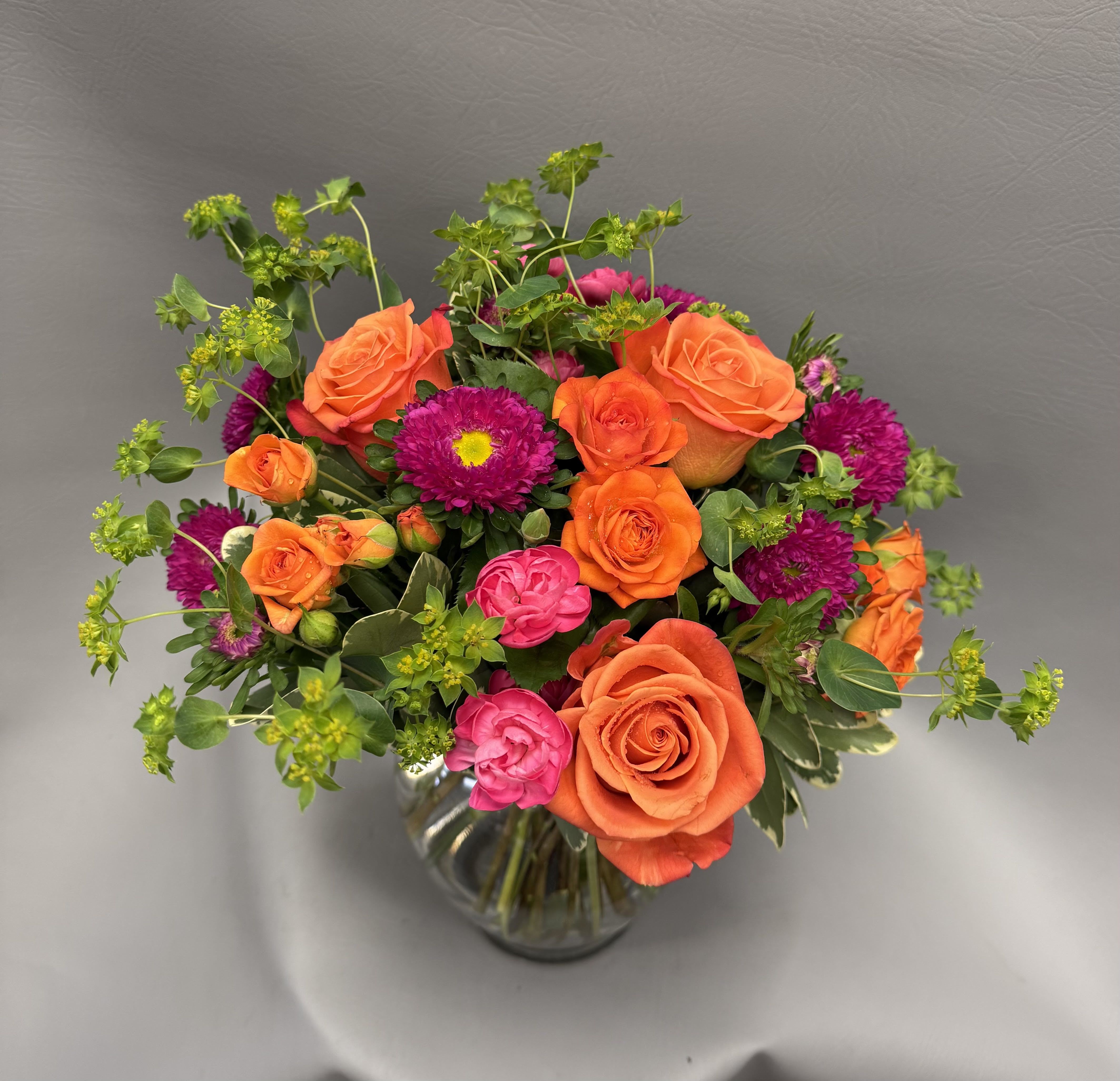Bright arrangement of orange roses, pink blooms, and purple asters in a clear glass vase