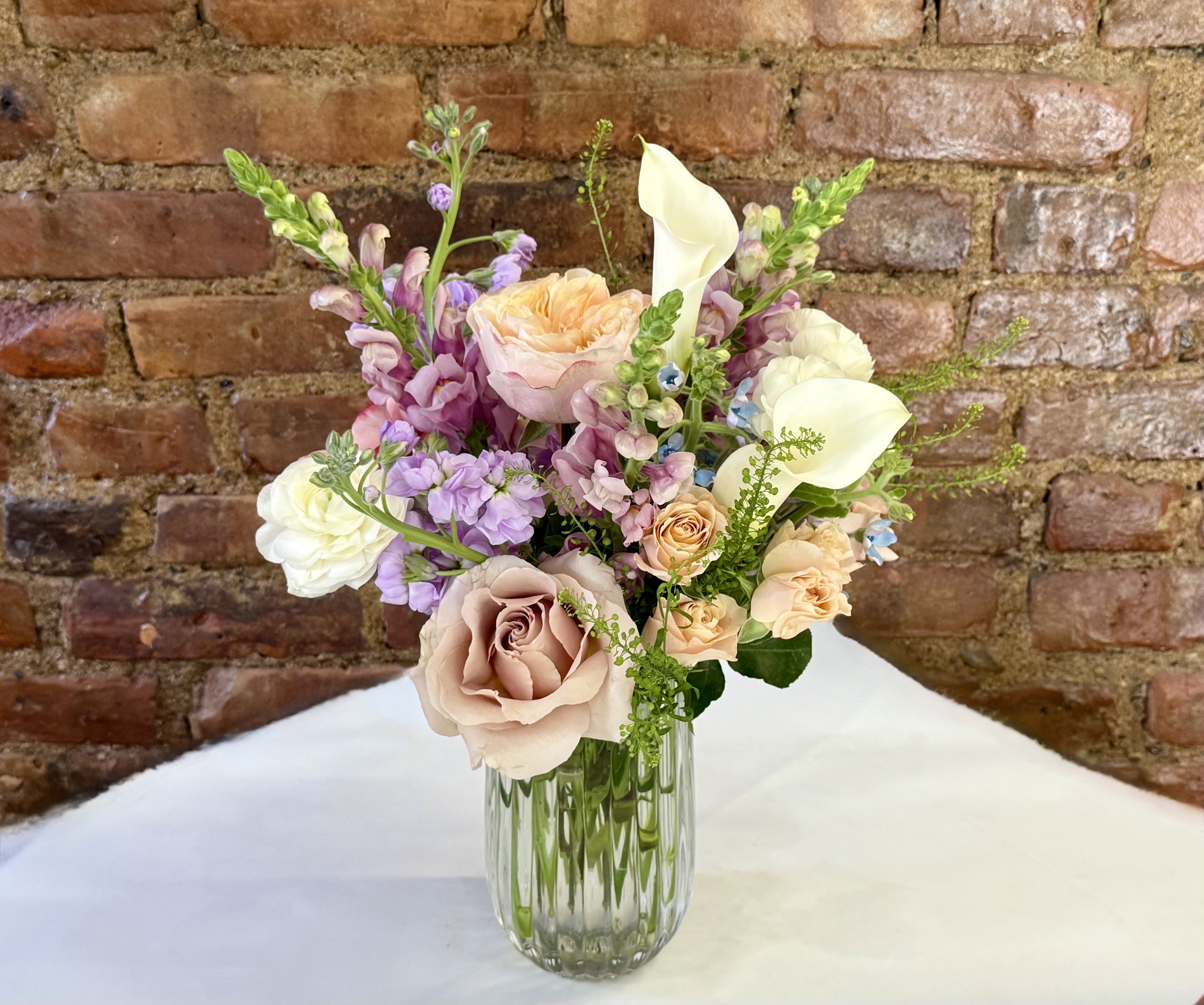 Pastel bouquet of roses, calla lilies, and purple blooms in a glass vase