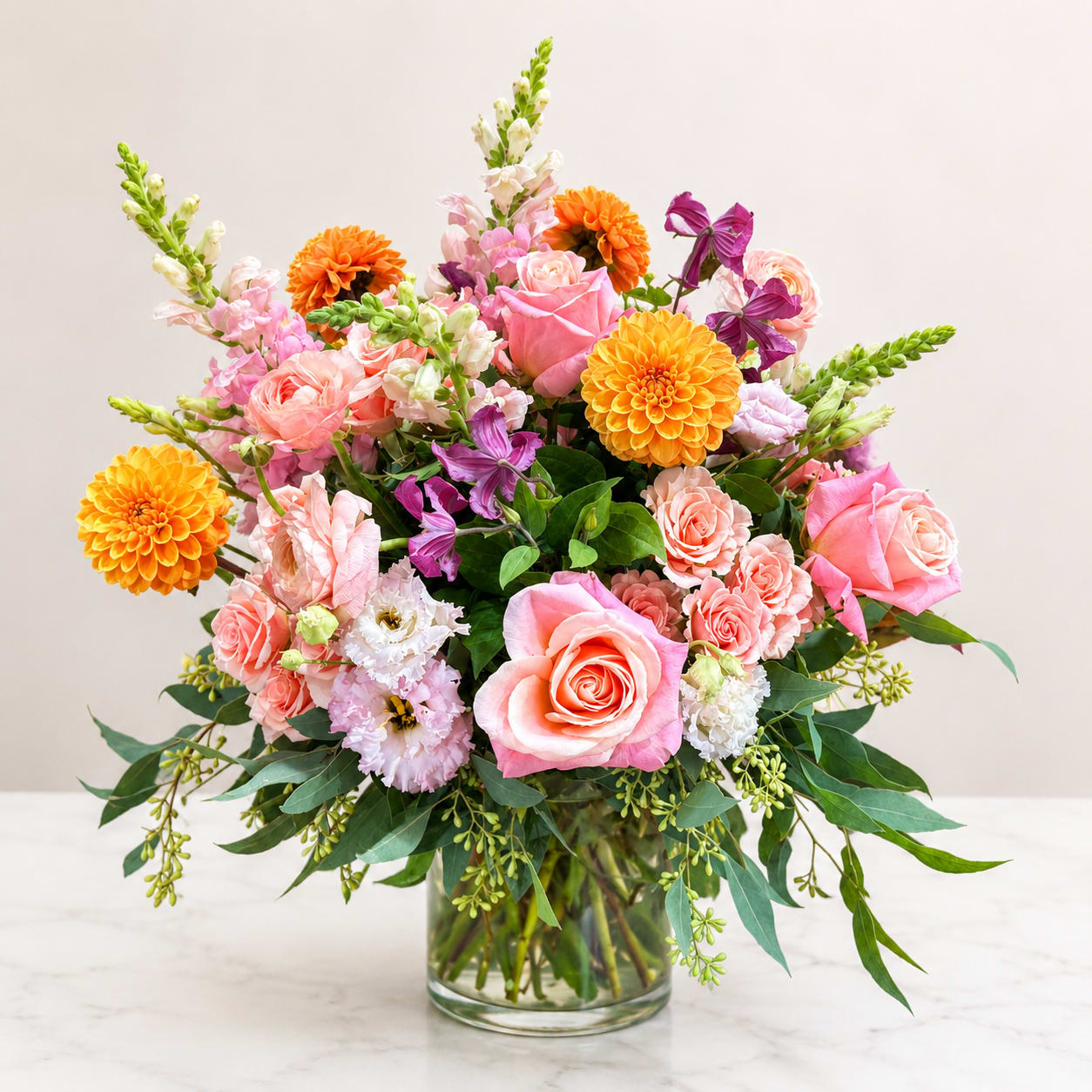 Sunset Garden - A vibrant garden-inspired arrangement featuring premium roses, dahlias, spray roses, snapdragons, lisianthus, clematis, and lush seasonal greenery, thoughtfully designed in a modern glass vase. Bursting with cheerful peach, blush, coral, and sunset hues, this luxurious bouquet is perfect for birthdays, celebrations, anniversaries, or simply brightening someone's day.