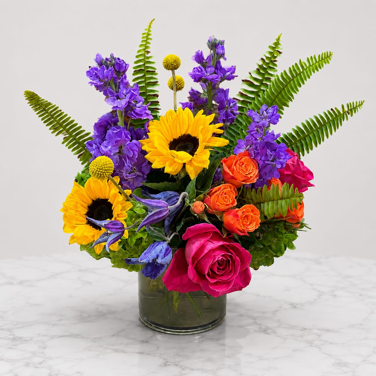 Sunshine Celebration - A joyful arrangement featuring radiant sunflowers, premium roses, vibrant stock, spray roses, clematis, craspedia, and lush seasonal greenery, artfully designed in a modern glass cylinder vase. Bursting with brilliant yellows, vibrant pinks, rich purples, and warm coral tones, this cheerful bouquet is the perfect way to celebrate life's happiest moments.