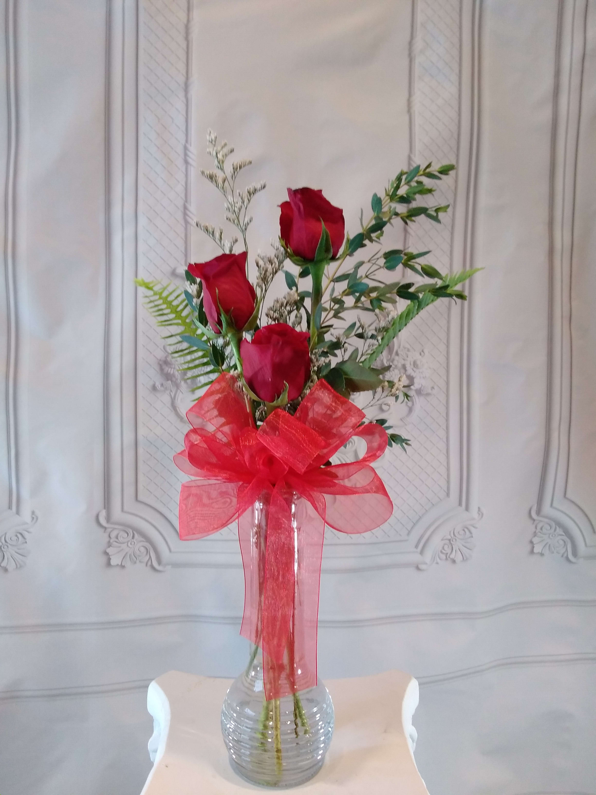 Three red roses in a clear glass vase with a large red bow