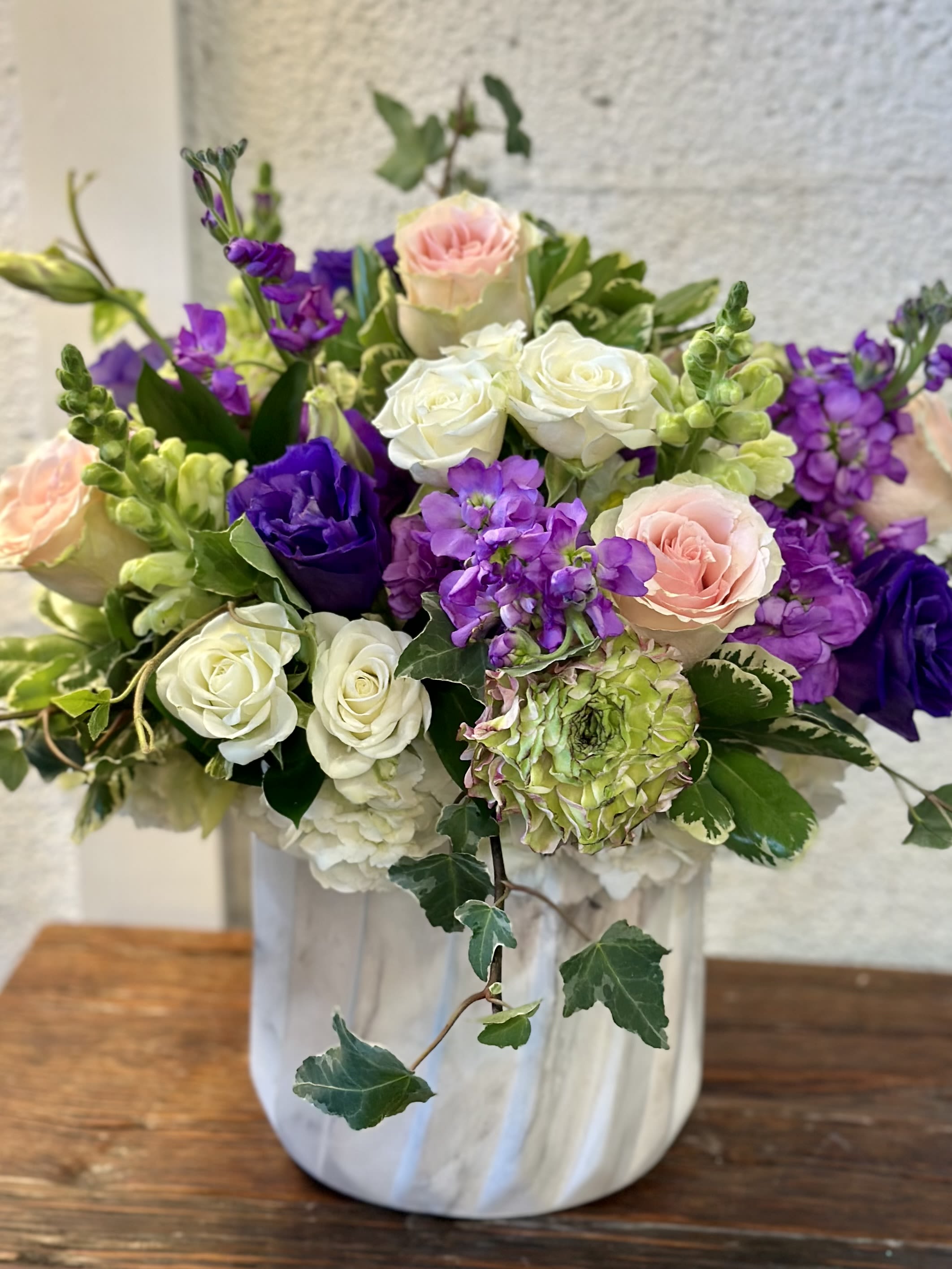 Pure Botanics  - Purple Lisianthus,  white, pink roses with hydrangea, stock and trailing ivy in a white ceramic vase - a lush, garden-style arrangement perfect for celebrations or a thoughtful everyday gift.
