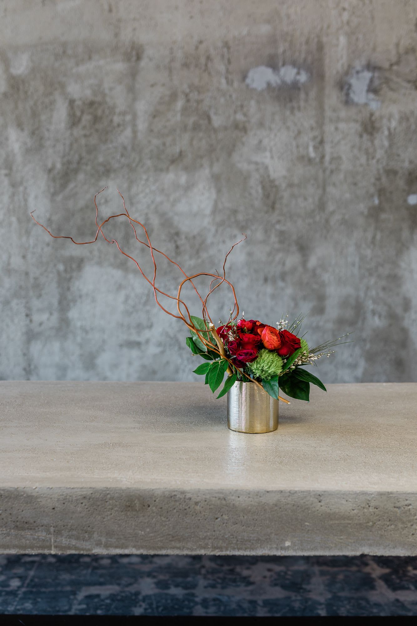 Red and orange floral arrangement in a metal vase with curly branches