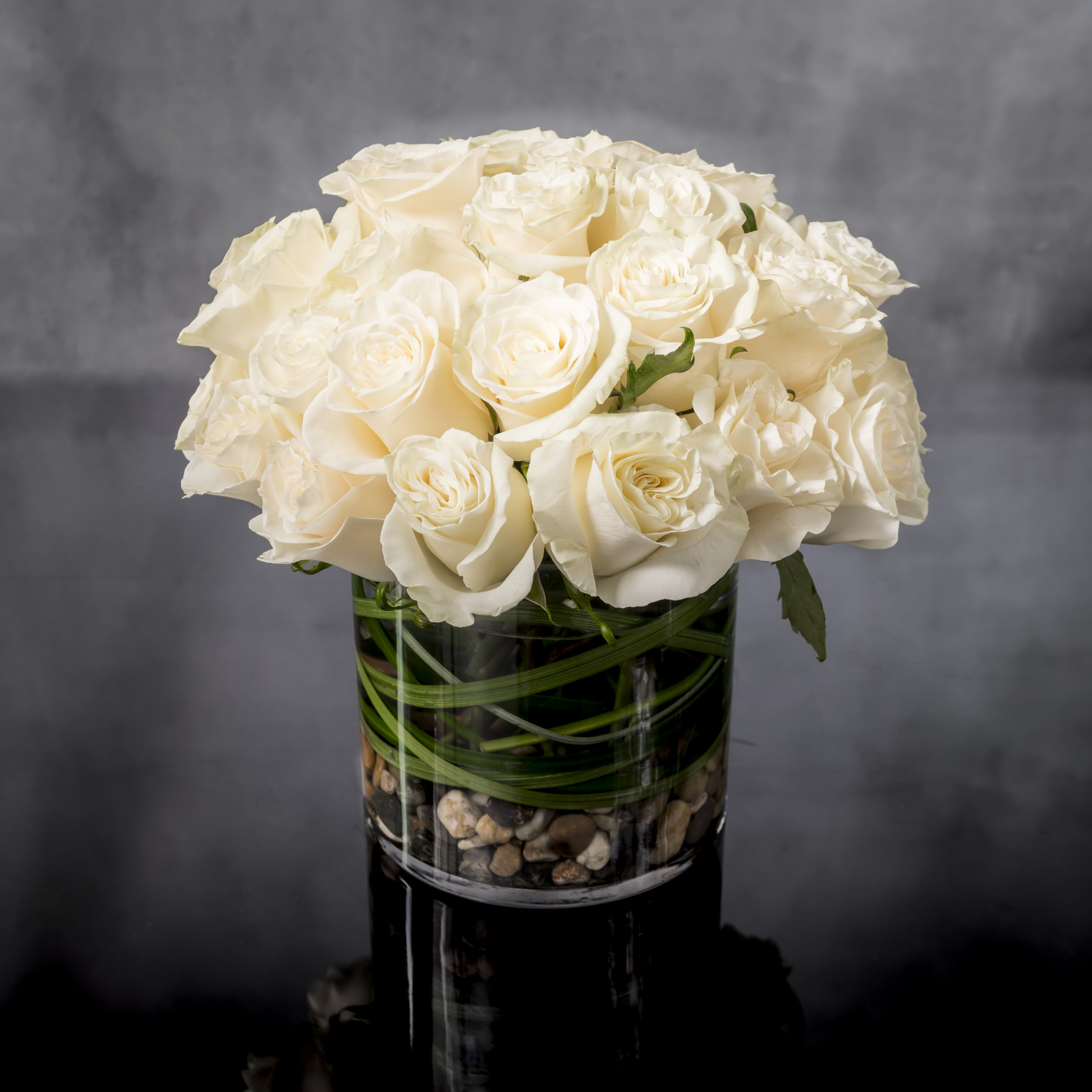 Compact arrangement of white roses in a clear glass cylinder vase with stones at the base