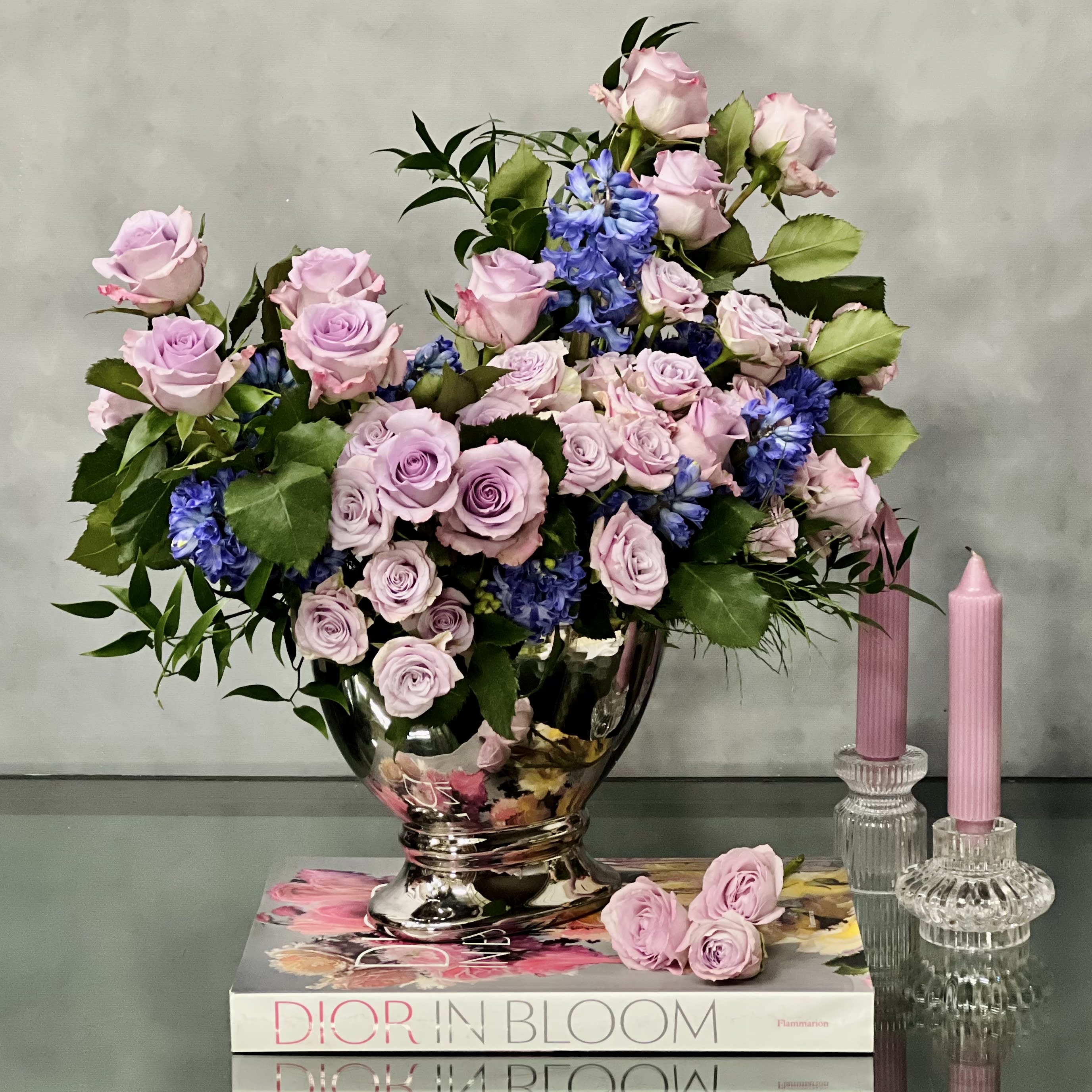 Lavender and pink rose arrangement with blue flowers in a silver vase beside pink candles