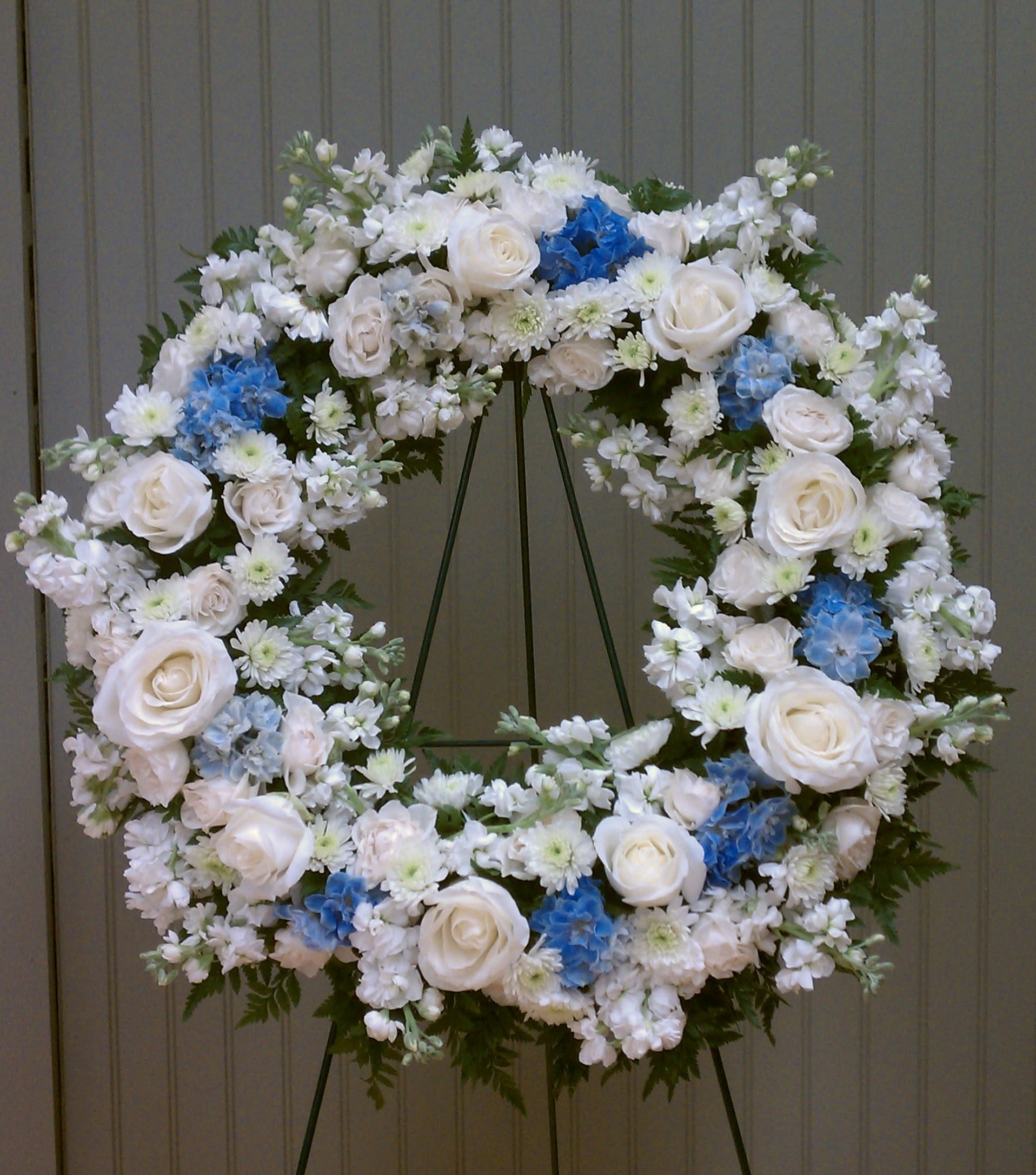 Large standing wreath of white roses and mixed white flowers with blue accents on a metal easel.
