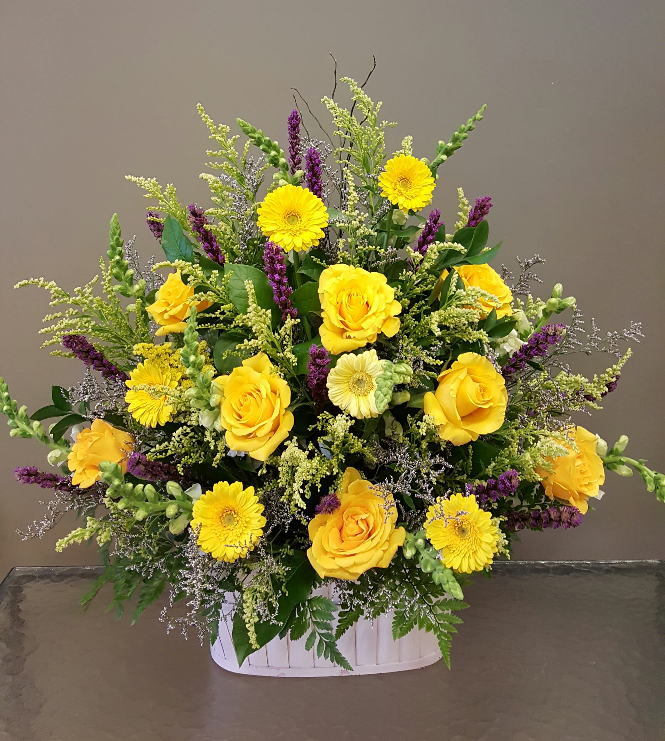Large yellow rose and gerbera daisy arrangement with purple accents in a white basket.