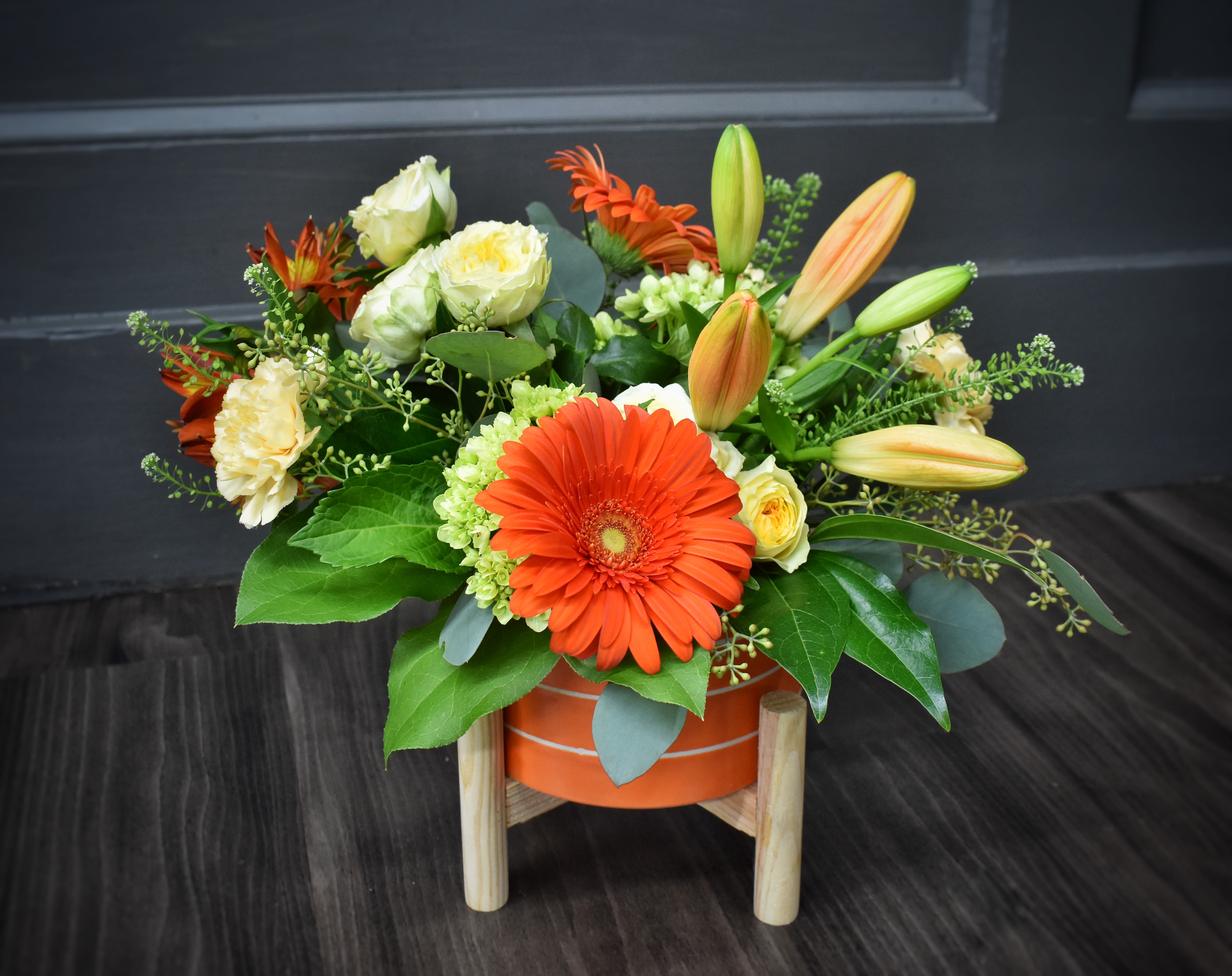 Sizzlin' Summer Blooms - A bold orange gerbera takes center stage in this Sizzlin' Summer Blooms flower arrangement, a birthday or congratulations bouquet that immediately reads as joyful and celebratory. The warm, citrus-forward palette - orange lilies and gerberas paired with yellow spray roses and pops of purple stock - makes the recipient feel noticed and genuinely cheered, perfect for birthdays, promotions, or "just because" moments.  Low and spreading with layered texture, the design mixes open blooms and unopened lily buds for movement, plus glossy green foliage and clustered green filler for contrast. You'll find gerbera daisies, lilies, roses, stock, carnations, hydrangea accents and other seasonal blooms arranged in a round orange container that sits on a simple wooden stand - a casual-but-polished presentation that's ready to display.  