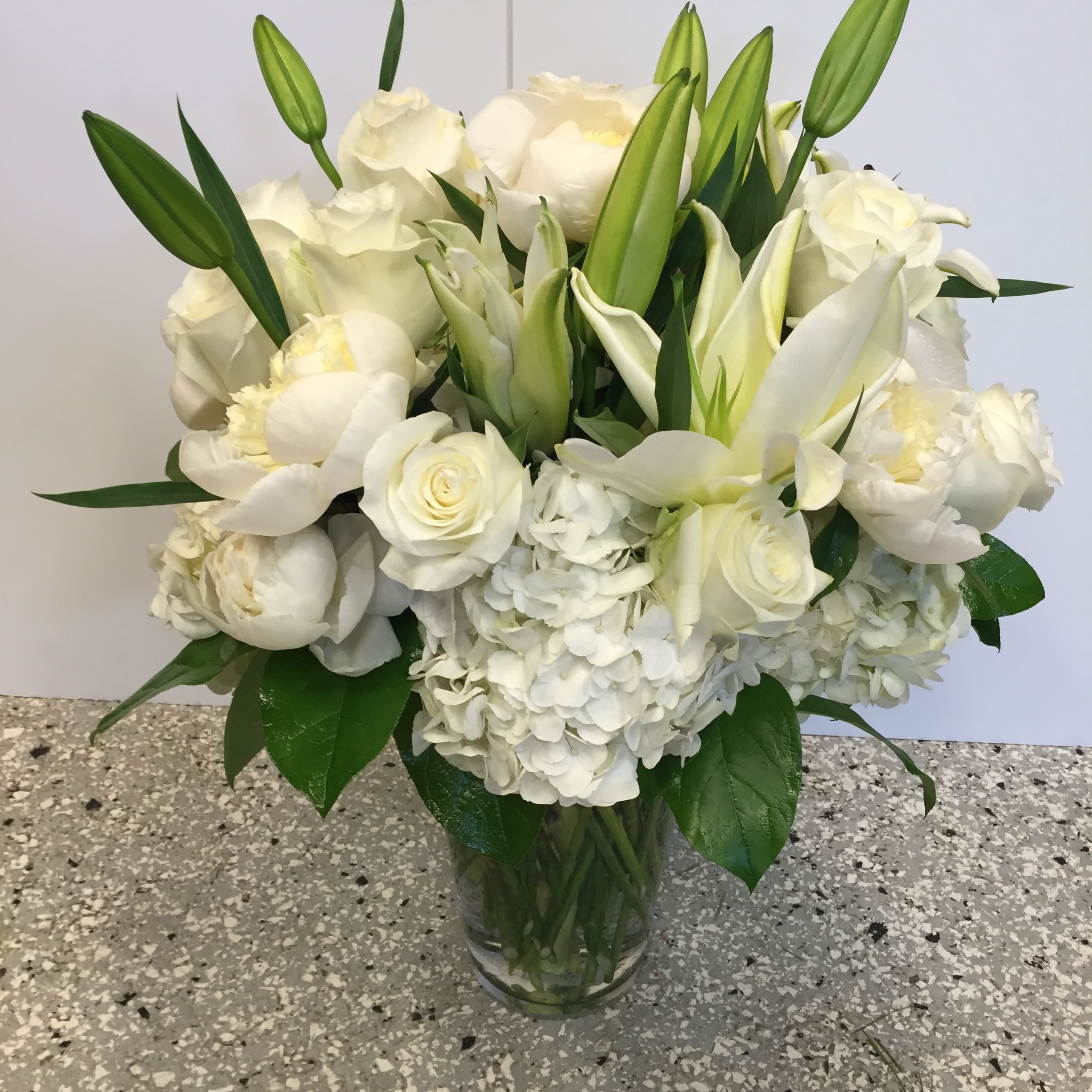 White floral bouquet in a clear glass vase with lilies, roses, and hydrangeas