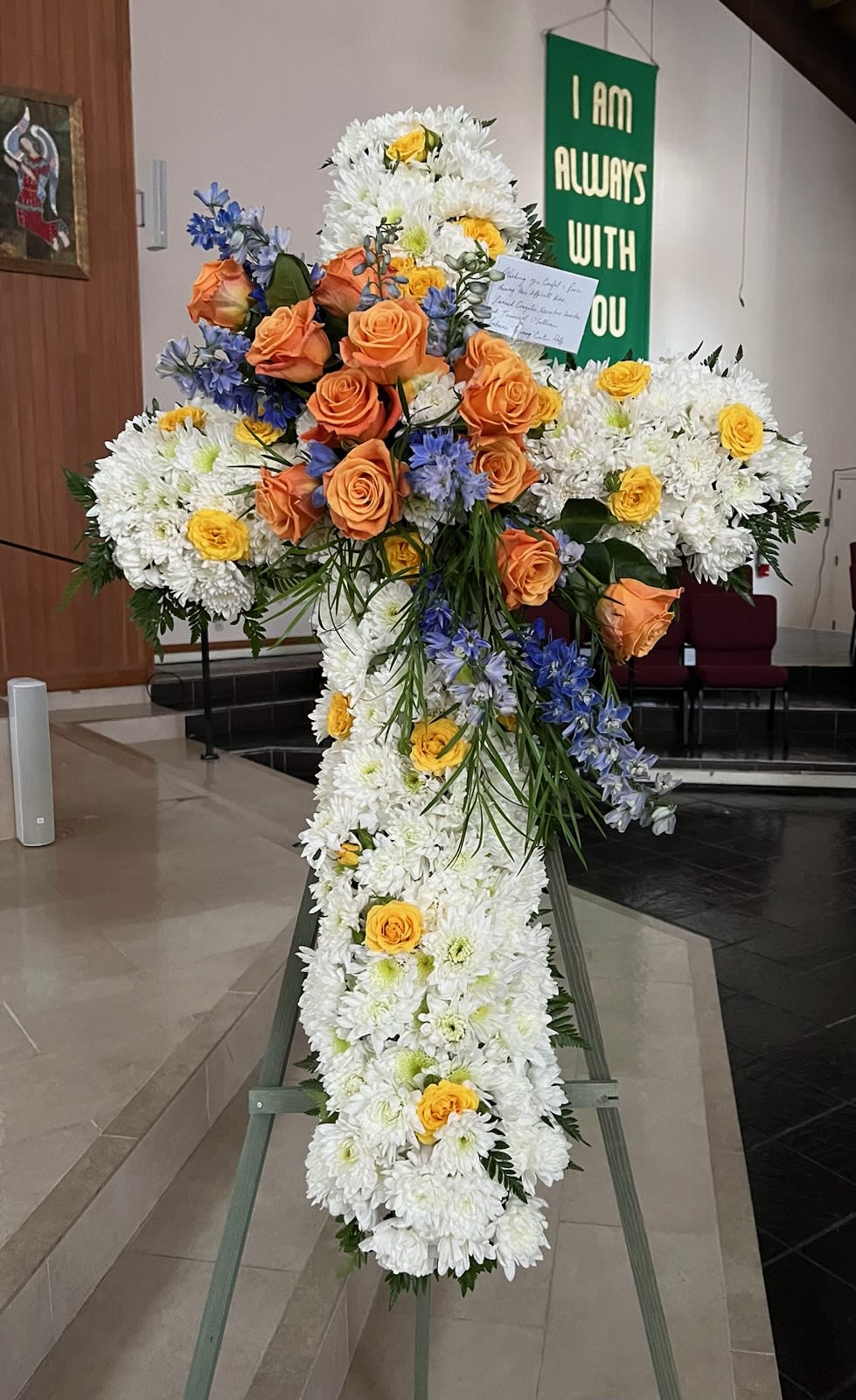 Large floral cross on an easel with white chrysanthemums, orange roses, and blue flowers