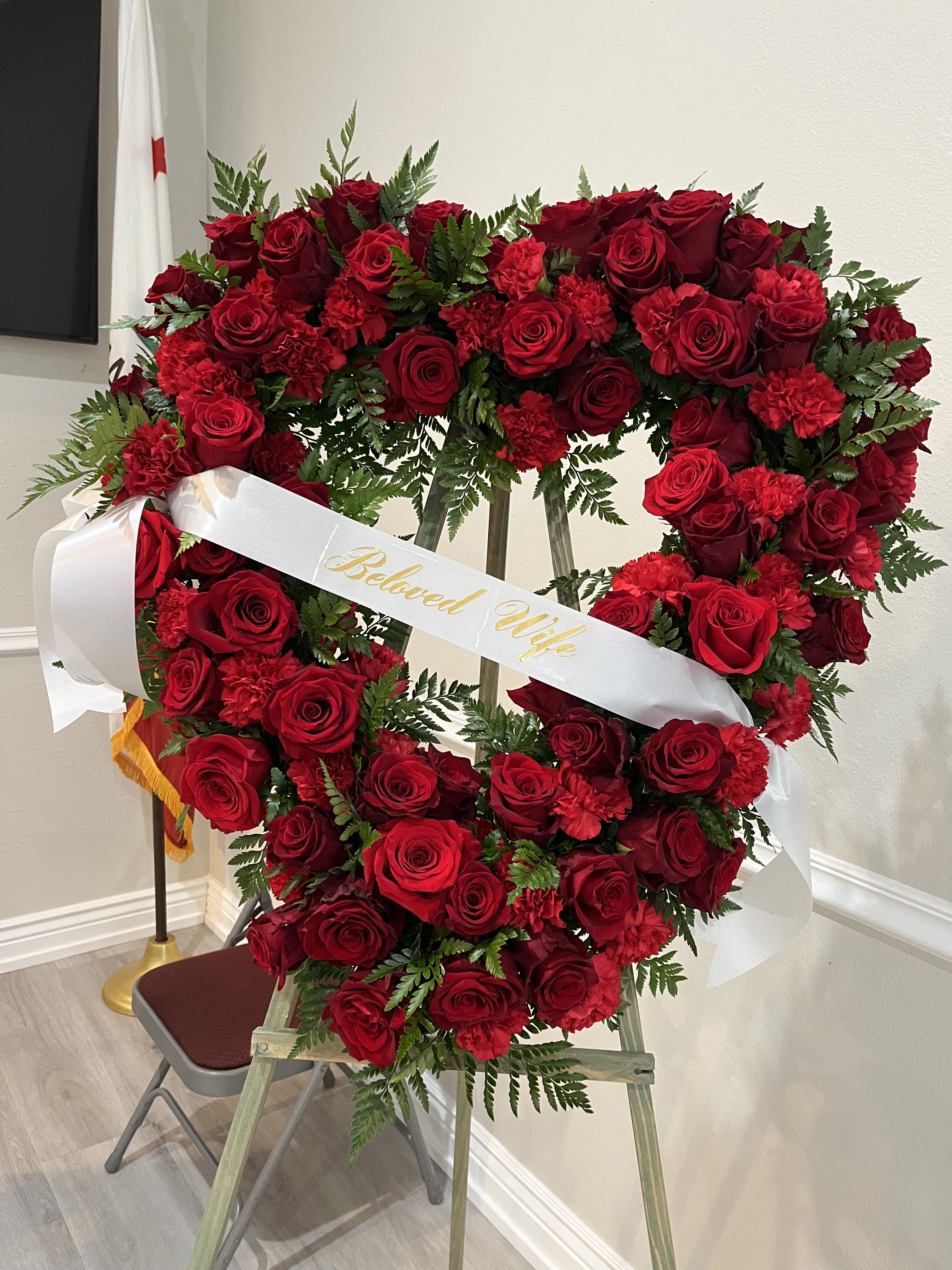 Heart-shaped red rose wreath with a white ribbon reading "Beloved Wife"
