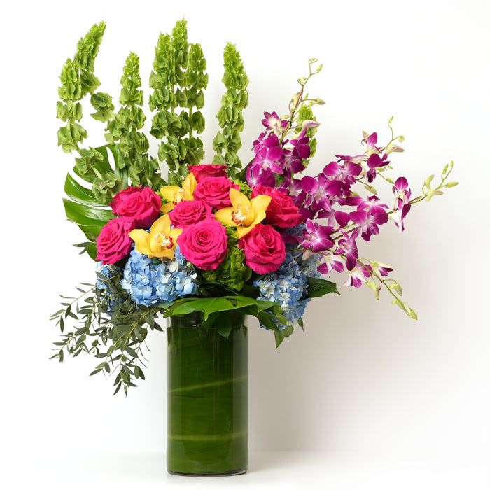 Tall arrangement of pink roses, orchids, and blue hydrangeas in a green-lined glass cylinder vase