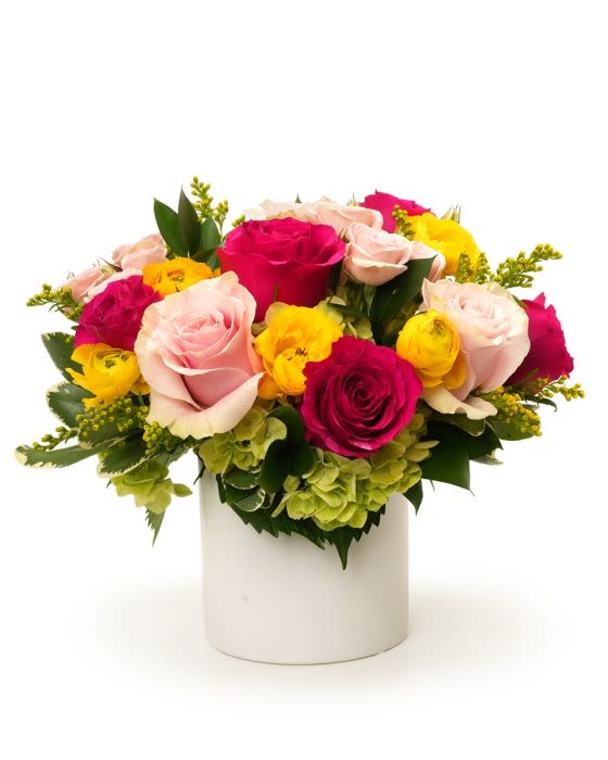 Low arrangement of pink and hot pink roses with yellow blooms and green hydrangea in a white cylinder vase