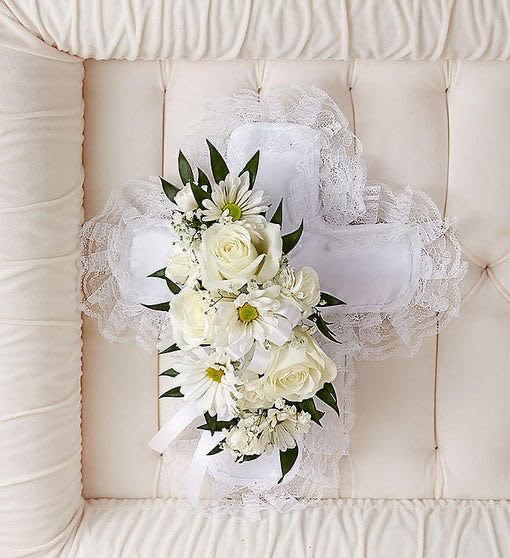 White floral cross pillow with roses and daisies resting on a cream tufted casket interior.