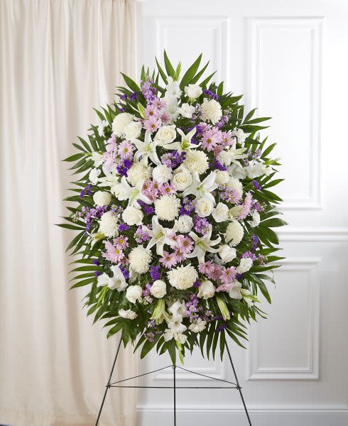 Tall standing spray of white lilies and roses with pink and purple flowers framed by green leaves on an easel.
