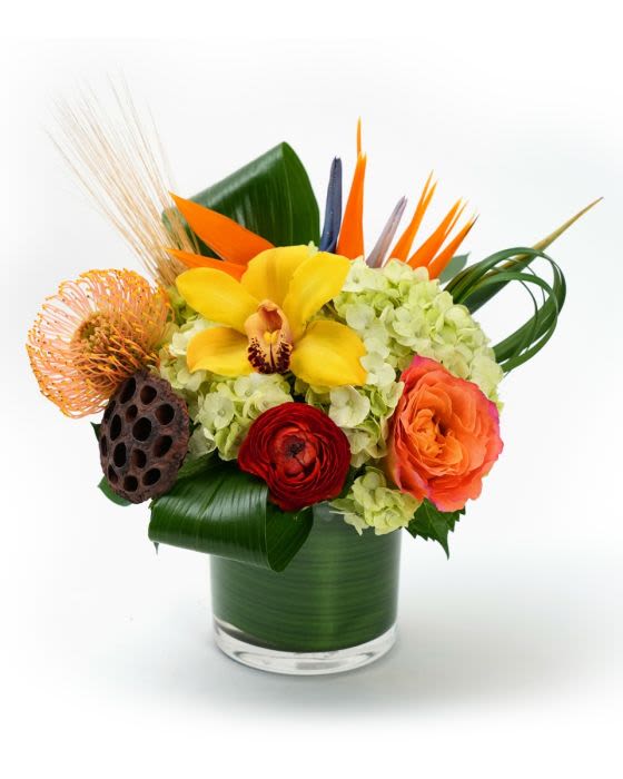 Colorful tropical arrangement with hydrangeas, orchid, rose, and protea in a leaf-wrapped glass vase