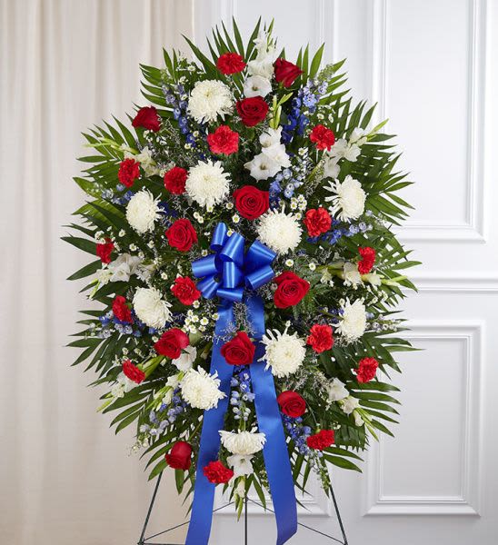Patriotic standing spray with red roses, white mums, blue delphinium and a large blue ribbon on an easel.