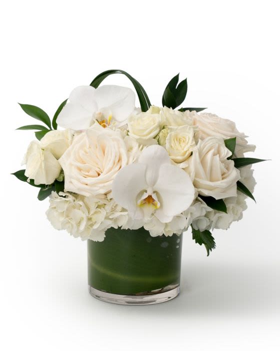 Compact arrangement of white roses, hydrangeas, and orchids in a low glass vase with green lining